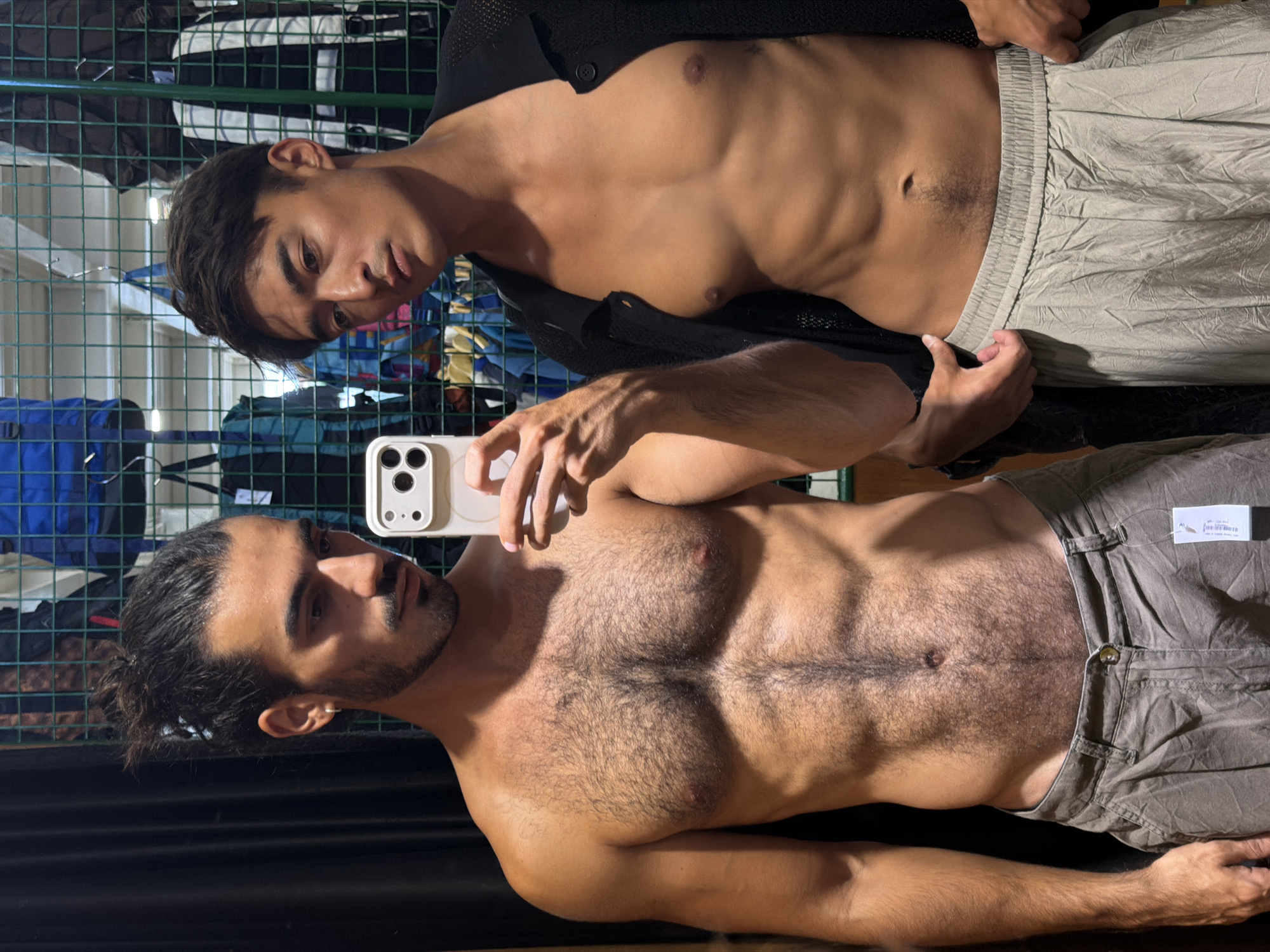 Two shirtless men are posing for a picture in a store. The man on the bottom has dark hair pulled back in a bun and is holding a phone up to take a selfie. He has a hairy chest and is wearing tan pants. The man on the top has dark hair and is wearing a black cardigan. He is also wearing tan pants. There are clothes racks and clothing behind them. The location is North Kuta, Indonesia.