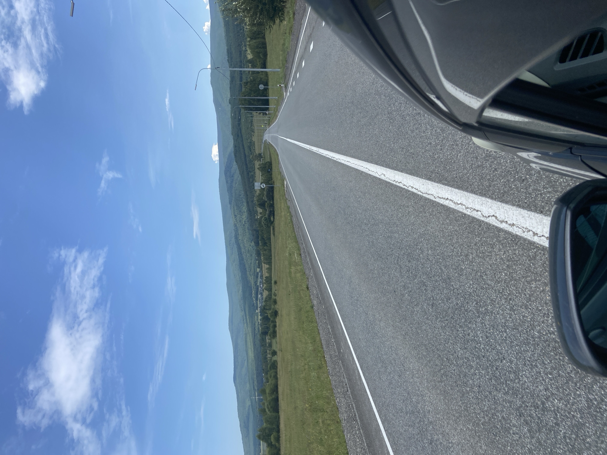 A wide asphalt road stretches into the distance, lined with a white dashed line. On the left, a grassy field meets a line of trees and rolling, tree-covered hills in the background. A blue sky with scattered white clouds covers the scene. The right side of the image shows the interior of a dark-colored car, including a side mirror and a portion of the hood. Streetlights line the road, and a few signs are visible. The scene appears to be a scenic route in a rural area, possibly near Zaktui, Russia, given the natural landscape and the presence of what seem to be typical Russian road features. There are no people visible in the image.