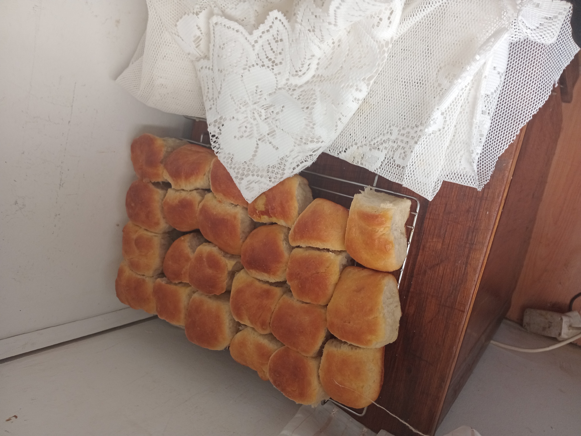 The image shows a metal cooling rack filled with freshly baked bread rolls. The rolls are golden brown and appear to be soft. They are arranged in rows on the rack. To the right of the cooling rack, a white lace tablecloth is draped, partially obscuring the background. The cooling rack rests on a wooden surface, possibly a table or countertop. In the background, a light-colored wall is visible, with a corner where two sections of the wall meet. A white electrical cord with a plug is visible on the floor in the lower left corner, near the wooden surface. The scene appears to be indoors. The image does not provide information about the specific location within Witzenberg Local Municipality, South Africa.