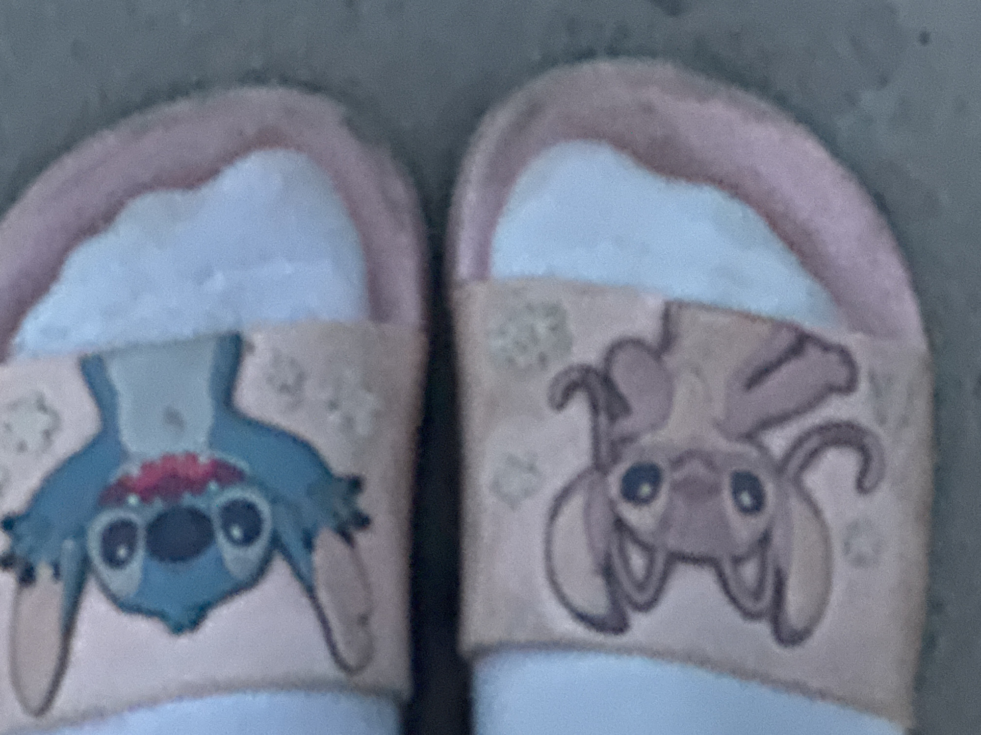 The image shows a pair of pink and white cartoon character slide sandals. The top sandal features a blue cartoon character with large eyes and a red mouth. The bottom sandal shows a light pink cartoon character with large eyes and antennae-like ears. The sandals appear to be on a paved surface, possibly a sidewalk or patio, in an outdoor environment. The location is described as Гожули, Ukraine. The overall impression is of casual footwear with a playful design.