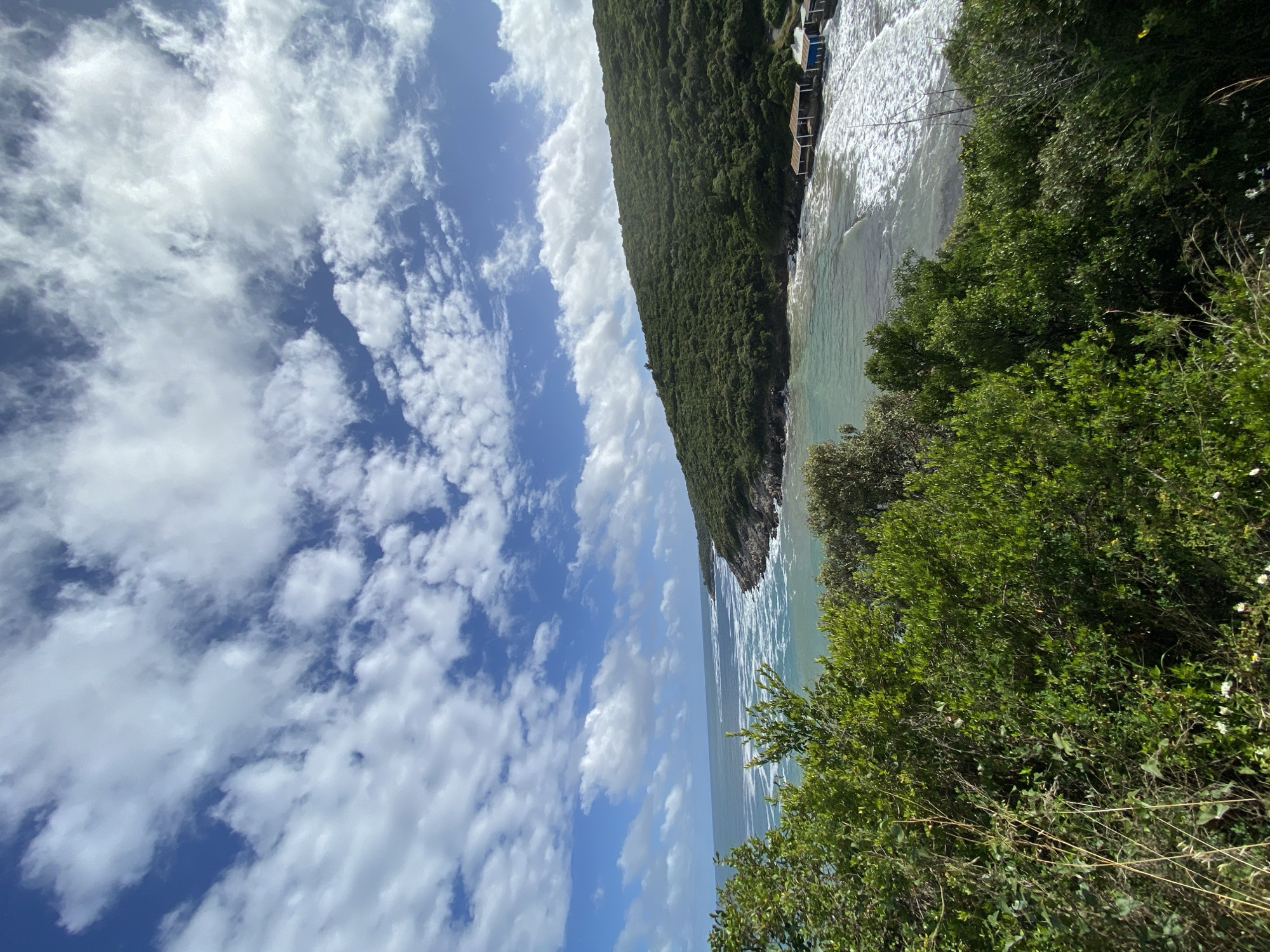 The image shows a coastal scene in Dubraljevina, Montenegro. A forested hill dominates the right side of the frame, sloping down to meet the sea. Buildings are visible on the far edge of the hillside. The water is light turquoise and appears choppy with white waves. On the foreground right, there's lush green vegetation, which obscures part of the scene. The left side of the image is dominated by a sky filled with puffy white clouds and patches of blue. The orientation of the image is slightly off-kilter.