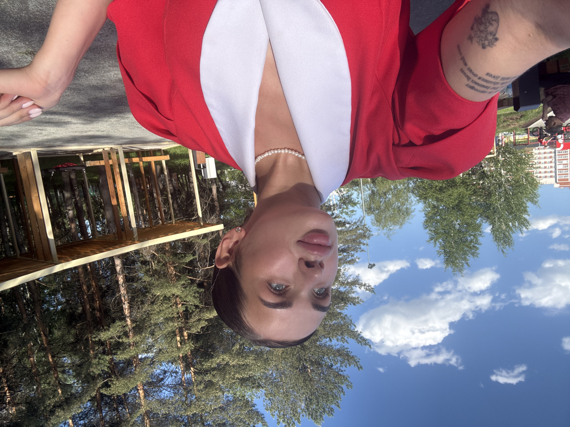 A woman is seen in an upside-down, waist-up portrait against an outdoor backdrop. She wears a red jacket with a white collar and a pearl necklace. She has light skin, dark hair, and blue eyes. Her right arm has a tattoo. The background includes green trees, a bright blue sky with white clouds, and a wooden structure that appears to be a gazebo. In the distance, an apartment building can be seen. The location appears to be a park or recreation area in Perm, Russia. In the background, another person wearing a red and white plaid shirt is visible.