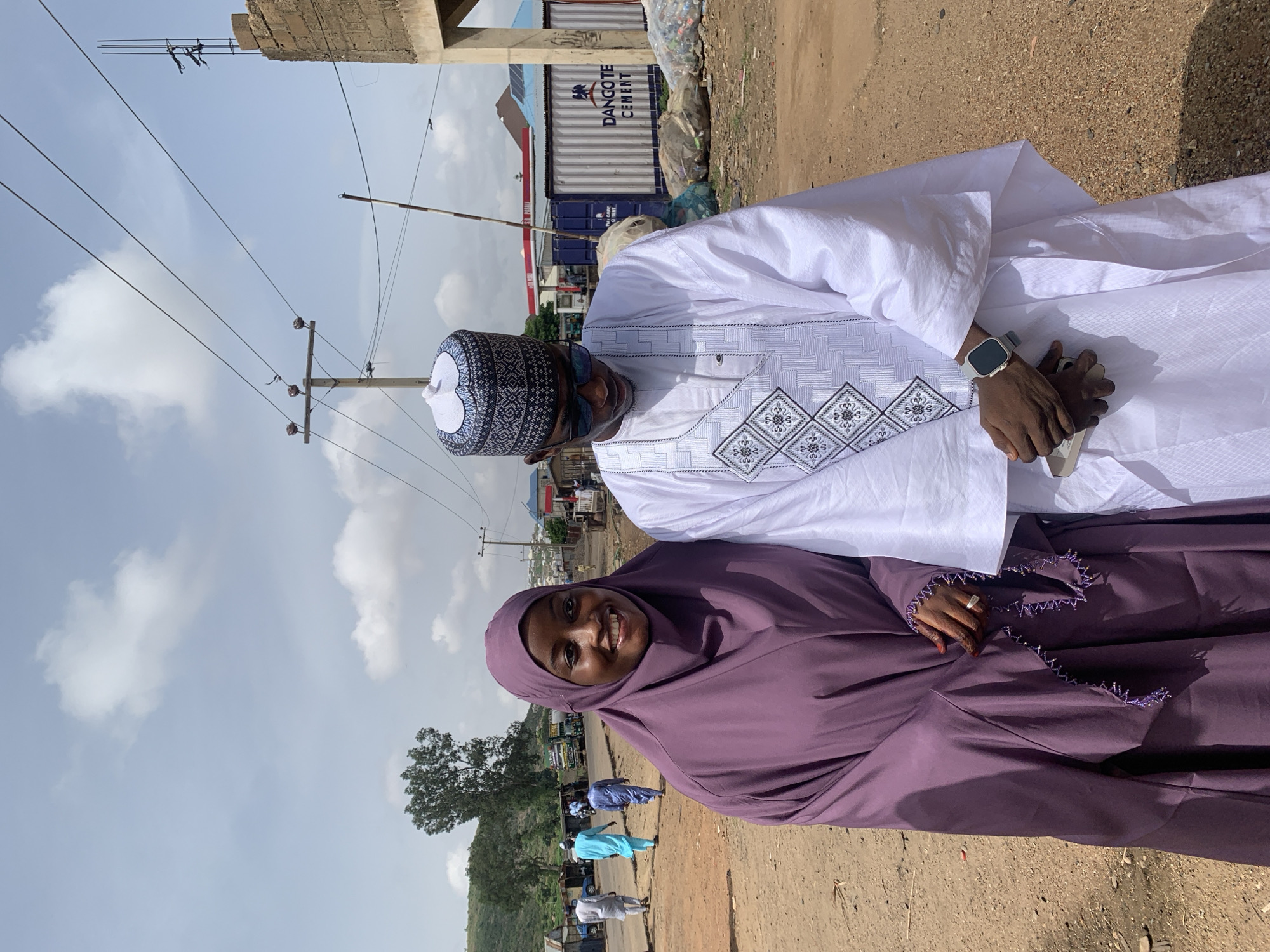 Two individuals are standing close to each other outdoors in Naraguta Hausawa, Nigeria. A woman on the left wears a purple hijab. A man on the right is in white attire, including a patterned hat. He is holding a cell phone and wearing a wristwatch.
In the background, there are utility poles with wires, a building, and shipping containers labeled "DANGOTE CEMENT." Other people are walking in the background. The ground appears to be dirt.