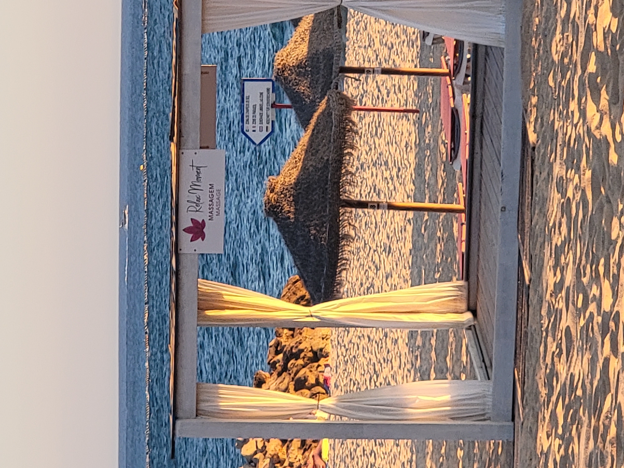 This is a photograph taken in Quarteira, Portugal, at a beachside massage establishment. The foreground shows a white wooden structure with sheer, cream-colored curtains hanging on either side, framing a view of the beach and the ocean. Beneath the structure, several lounge chairs with orange cushions are visible on a wooden deck. Two thatched umbrellas provide shade, with a wooden pole supporting one.
In the background, the calm blue ocean stretches to the horizon under a clear, pale sky. A thin line of buoys is visible in the water, and a small white boat can be seen in the distance. To the left of the frame, a rocky outcrop juts into the sea. A sign on the white structure reads "Relax Moment" with a stylized pink lotus flower logo, and below it, "MASSAGEM MASSAGE". To the right of this sign, a directional arrow sign points left, with text in Portuguese, indicating services or facilities. The beach sand in the foreground shows the textured patterns left by the receding tide or wind. The lighting suggests it is late afternoon or early morning, with the sun casting warm, long shadows.