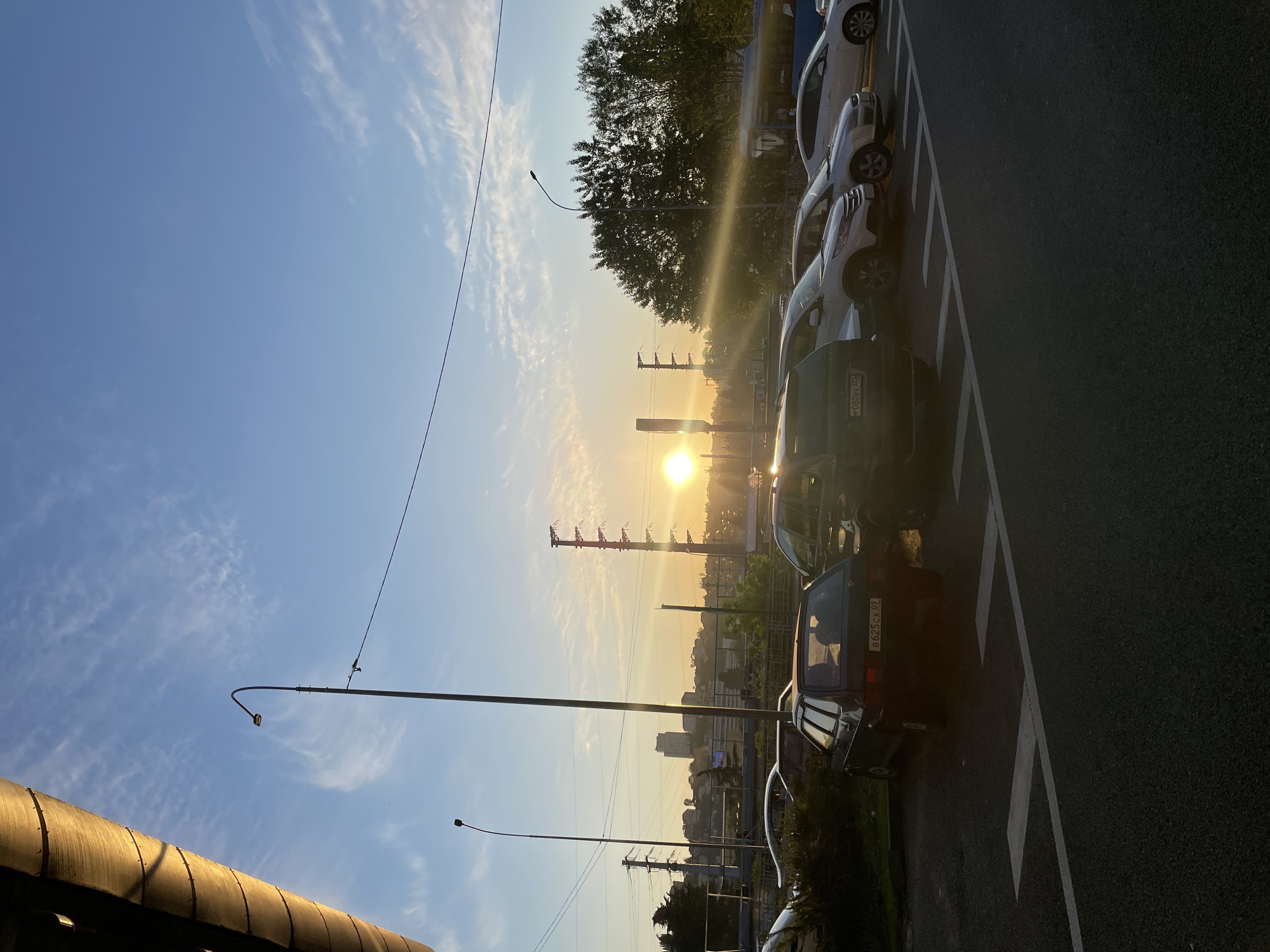 The image, taken in Sochi, Russia, depicts an urban street or parking lot scene during either sunrise or sunset, characterized by the bright, low sun on the right casting a strong golden glow and lens flare. The sky is clear blue with wispy, scattered white clouds.
Several cars are parked in a row on the right side of the frame, positioned on an asphalt surface marked with white diagonal lines, suggesting a parking area or roadside. The vehicles are mostly light-colored, with one prominent dark green/blue car in the foreground and a lighter car further back. License plates are visible on two vehicles: "Р 080 BK 23" on the dark car, indicating the Krasnodar Krai region (where Sochi is located), and "В 825 СХ 09" on a light-colored car, corresponding to the Karachay-Cherkessia region.
Multiple tall utility poles or streetlights with curved arms are silhouetted against the bright sky, along with visible power lines. In the mid-ground, a lush canopy of green trees is visible, backlit by the sun. Beyond the trees, the faint outlines of distant city buildings and a hazy horizon complete the urban landscape. A large, ribbed, metallic structure, possibly part of an overhead bridge or a building roof, occupies the bottom-left corner, reflecting the warm sunlight. The scene is quiet, with no explicit human activity, conveying a calm and serene atmosphere typical of the "golden hour."