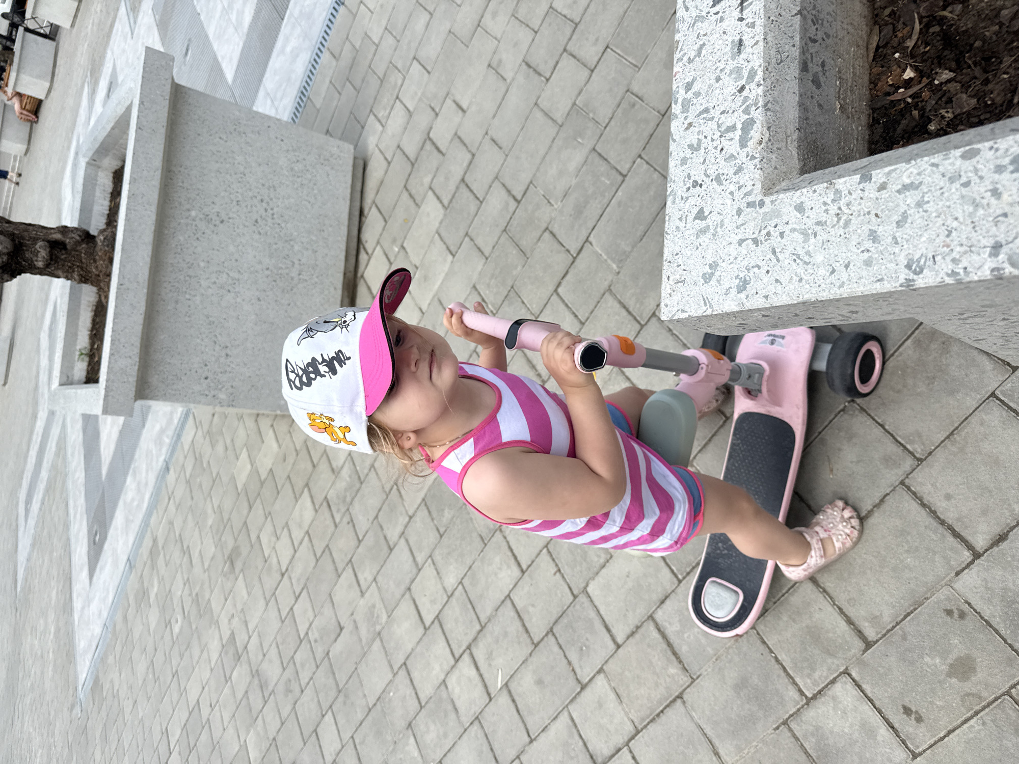 A young Caucasian girl with blonde hair is standing on a pink scooter outdoors. She is wearing a pink and white striped tank top, blue shorts with pink trim, and pink sandals. She is also wearing a white baseball cap with cartoon characters on it and a pink brim. The girl is holding onto the handlebars of the scooter and looking to her right. The scooter is pink and black with three wheels. The background is a paved courtyard with gray rectangular stones. There are large concrete planters with trees in the background. The lighting suggests it is daytime. The location is Unknown, Unknown.