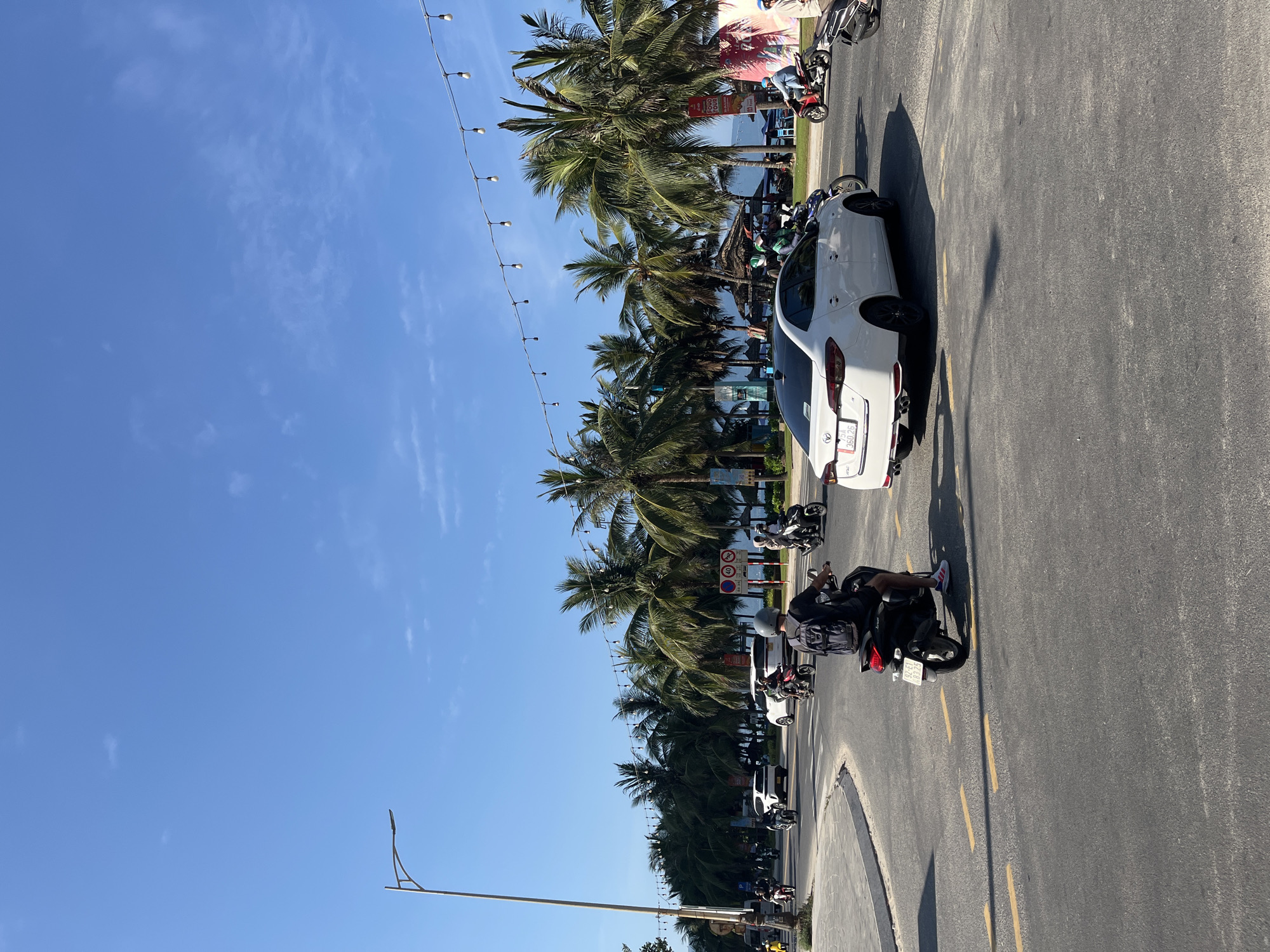 The image shows a street scene in Đà Nẵng, Vietnam. Palm trees line the street, suggesting a coastal location. The sky is clear and blue. A white car is parked on the side of the road. Several motorcycles are also present, some in motion and some parked. A string of lights is strung above the street. The road surface is asphalt and marked with yellow lines. People are on the motorbikes.