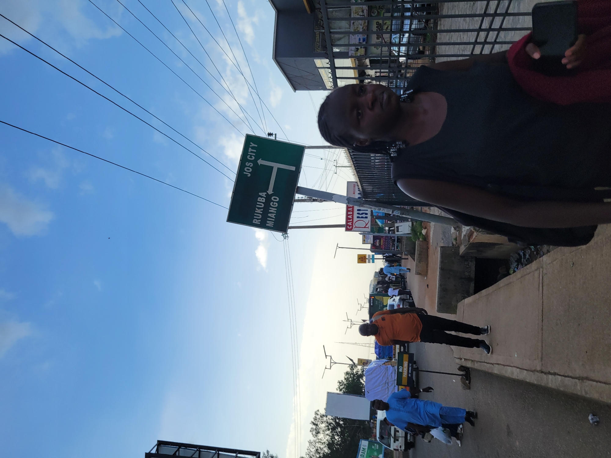 A busy street scene in Jos, Nigeria, captures a moment of urban life. A woman in a dark sleeveless top walks towards the viewer, her expression neutral, carrying a dark bag on her left shoulder and a red garment in her right hand, holding a phone. In the background, a green road sign indicates "JOS CITY" with an arrow pointing right, and "RUKUBA MIANGO" with a left-pointing arrow. Traffic, including buses and cars, is visible on the road, with pedestrians walking on the sidewalk. A man in an orange t-shirt and black trousers walks away from the viewer, while another man in a blue traditional outfit walks towards the viewer. Streetlights are present along the road. The environment appears to be a commercial area with buildings and a fence along the sidewalk. The sky is partly cloudy with a clear blue expanse.