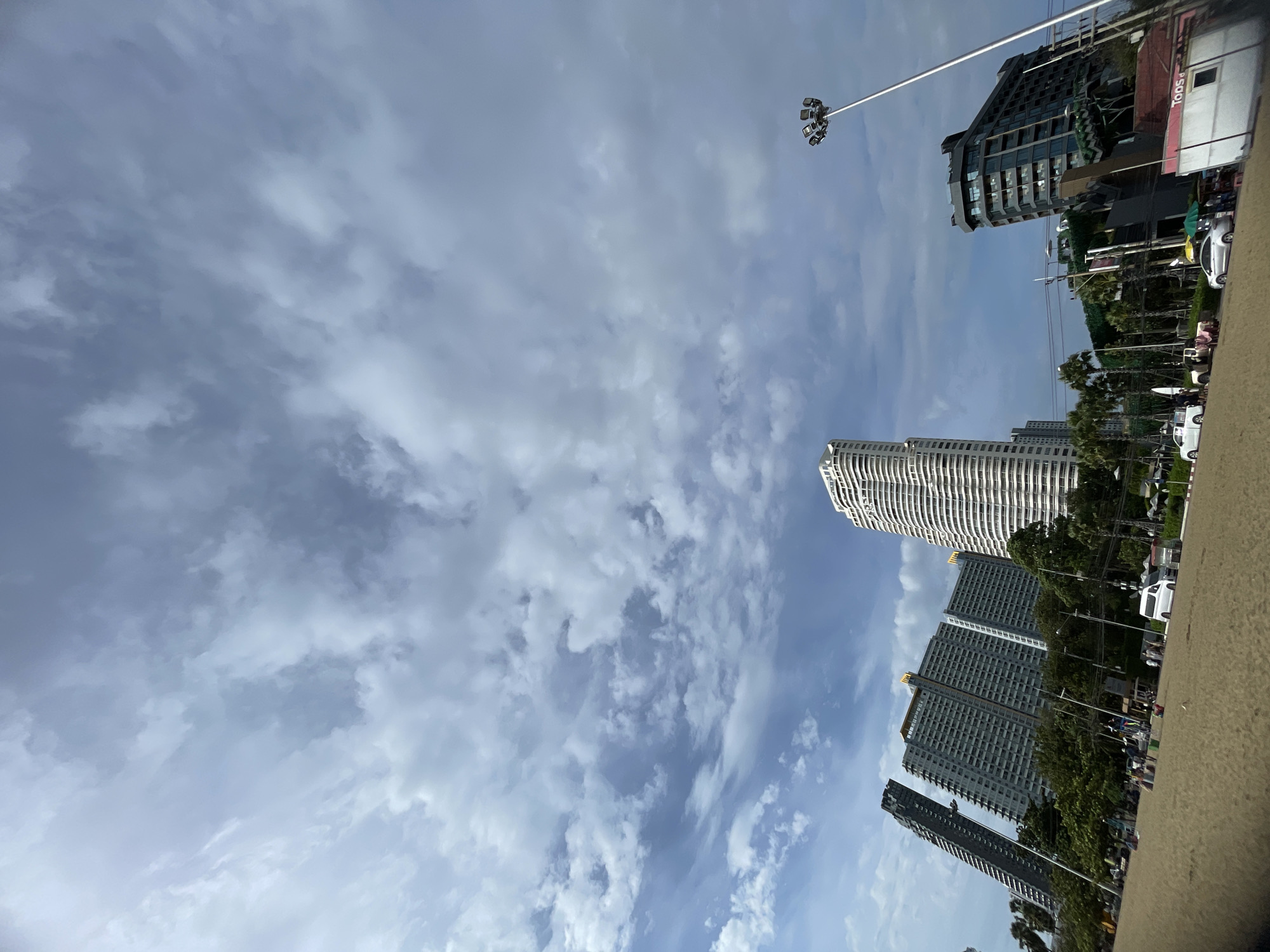 The image shows a beach scene in Pattaya City, Thailand, under a cloudy sky. Several tall buildings, including a distinctive white, spiraling tower and a dark, angular building, dominate the skyline. Palm trees and other vegetation line the area behind the beach. People are scattered across the sandy beach, some sitting or walking, while several white vehicles, including a van and cars, are parked on a paved area near the buildings. A tall lamppost with multiple lights is visible on the right side of the frame. The overall impression is a bustling urban beach environment.