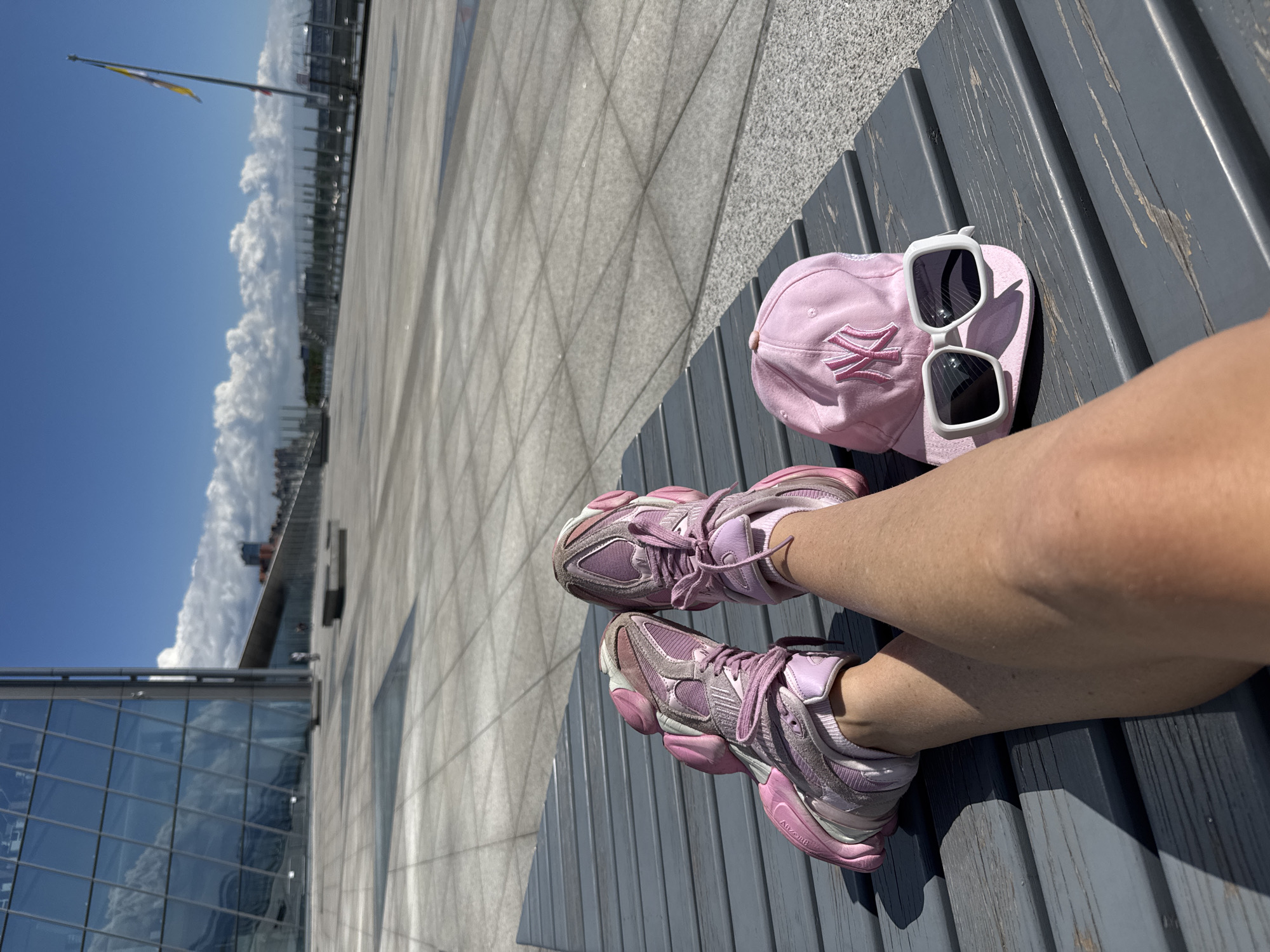 The image presents a first-person perspective of someone resting on a grey slatted bench in an outdoor urban environment, identified as Saint Petersburg, Russia. The subject's lower legs and feet, adorned with pink athletic sneakers, are central in the foreground. The sneakers feature a chunky design, with "ABZORB" visible on the sole. Adjacent on the bench are a pink baseball cap, embroidered with a white "NY" logo, and white-framed sunglasses with dark lenses.
The scene unfolds under a clear blue sky, marked by fluffy white cumulus clouds. Bright sunshine illuminates the area, casting distinct shadows and indicating it is daytime, likely mid-morning or afternoon with pleasant weather. In the background, a vast, light-colored paved expanse stretches towards modern buildings characterized by large glass panels and geometric architectural patterns. A tall flagpole, flying a flag with yellow and blue stripes, stands against the horizon. The overall impression is one of relaxation and a break amidst urban exploration or leisure activity.