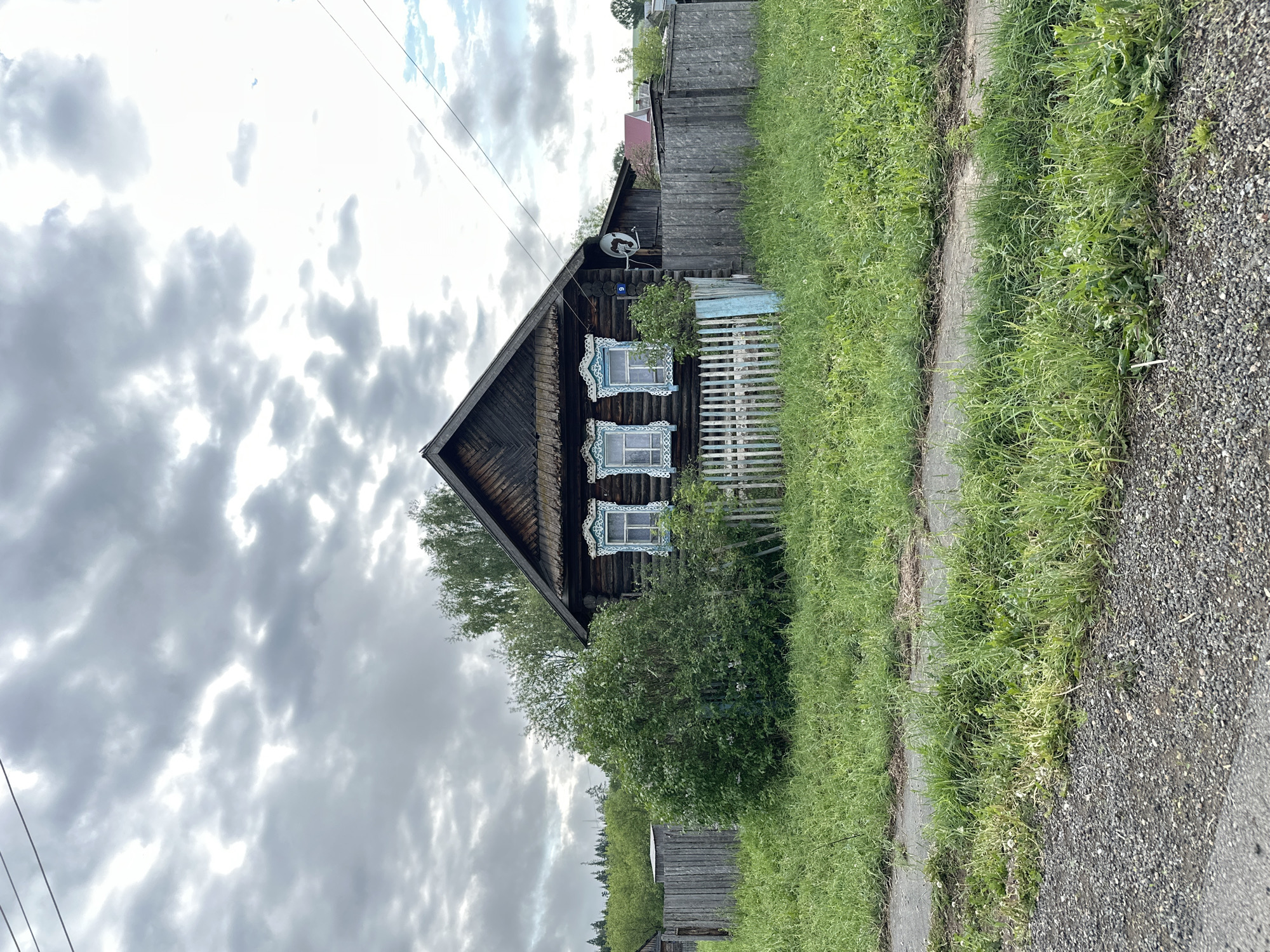 An image captures a quaint, weathered wooden house in the Russian village of Sredneye Kechevo. The house, with its distinctive triangular roof, features three windows adorned with intricately carved, light blue trim. A satellite dish is attached to the upper portion of the house. The structure is partially obscured by lush greenery, including a dense bush in front and overgrown grass along a paved walkway. A weathered wooden fence extends along the property line. The overcast sky suggests a cloudy day, and telephone wires cross above. A narrow gravel road borders the edge of the frame.