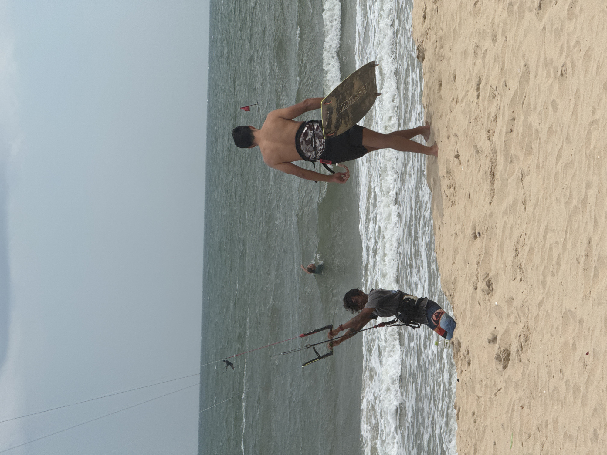 In Mũi Né, Vietnam, on a sandy beach adjacent to a choppy, gray ocean under an overcast sky, two men are engaged in kitesurfing activities. The foreground shows a man holding onto the control bar of a kite, positioned near the water's edge. He is wearing a dark t-shirt, dark shorts with colorful patches, and a kiteboarding harness. His left leg is bent, and he is looking towards the ocean. Behind him, further out in the water, another person appears to be swimming or in the process of kitesurfing. In the mid-ground, a second man, shirtless and wearing black shorts with a camouflage-patterned harness, walks away from the camera towards the ocean, carrying a yellow surfboard. A small red flag is visible on a pole in the ocean in the distance. The waves are breaking on the shore, creating white foam.