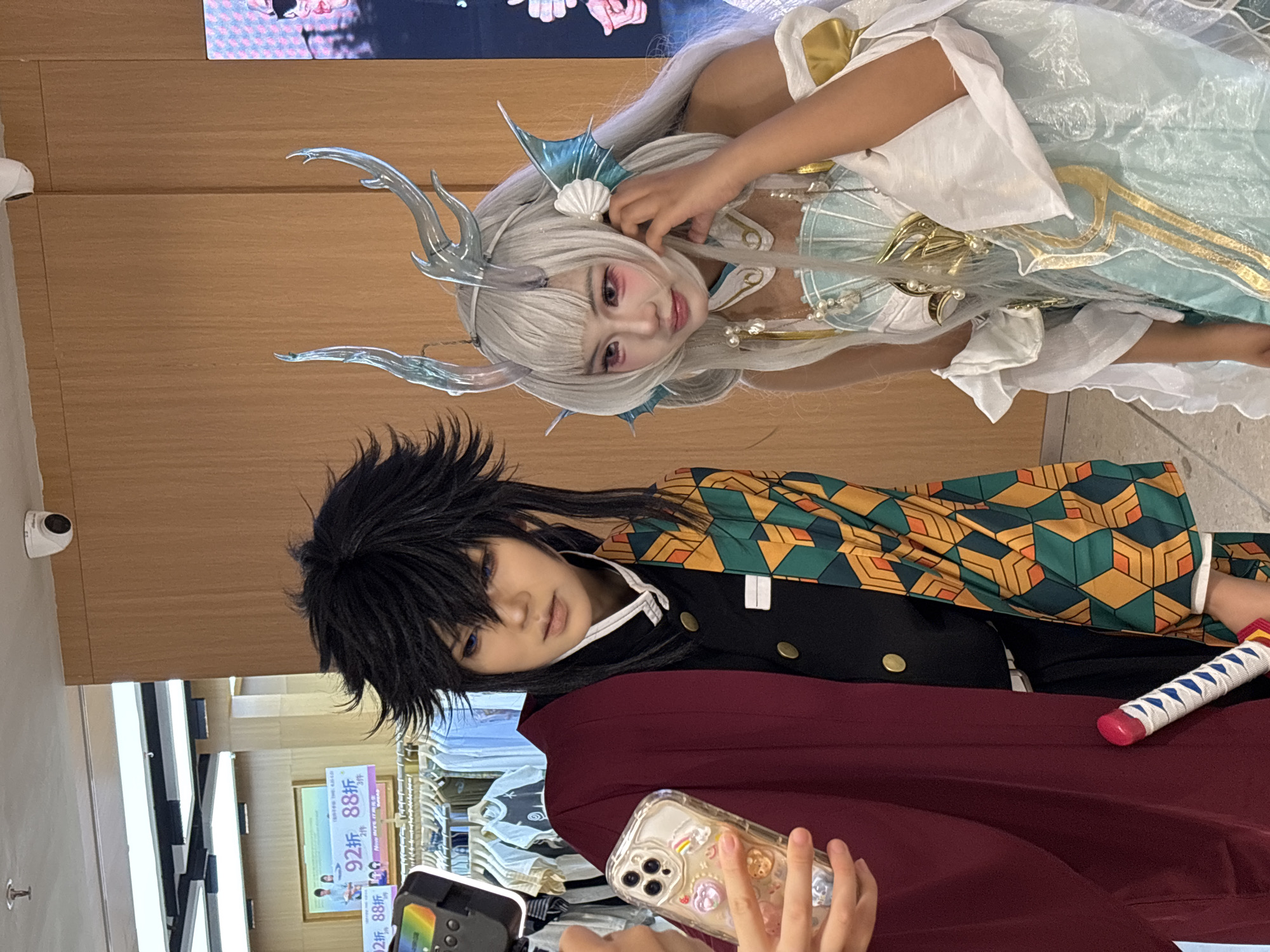 Two cosplayers are posing for a photo inside a store. One is dressed in a "Demon Slayer" outfit, with a black wig, kimono, and sword. The other is dressed in a fantasy outfit with a white wig, headpiece, and a flowing white and light blue dress with gold accents. An individual is holding a smartphone and a light in front of them. The setting appears to be a retail store, indicated by clothing racks and promotional signs. This is likely in the Liangjiang New Area of Chongqing, China.