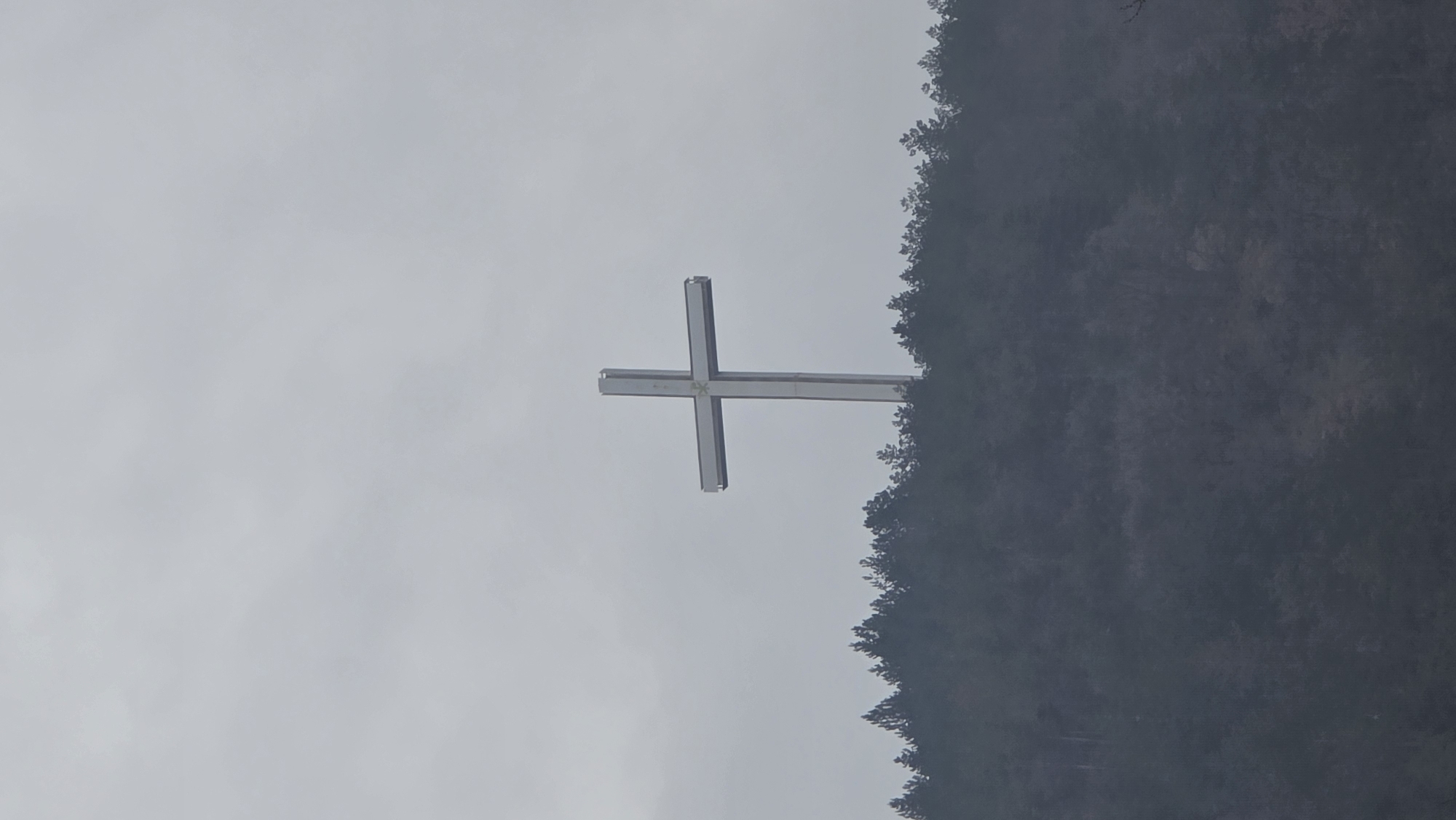 The image features a large metal cross against a gray sky. The cross is prominently positioned, appearing to stand on a hill or mountainside covered with trees. The location is Blagoevgrad, Bulgaria.
