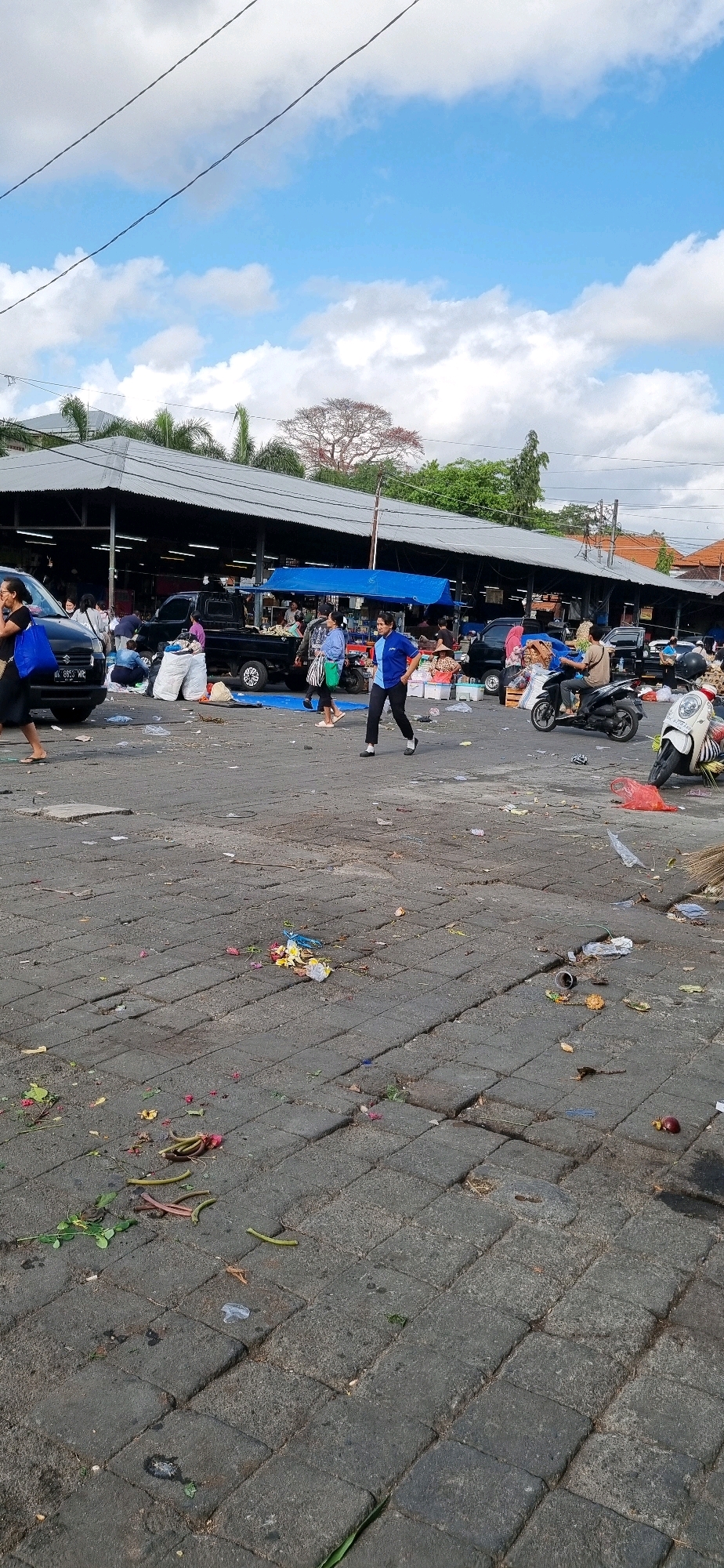 The image depicts a street scene, likely a market, in Denpasar, Indonesia. Paved ground, cluttered with debris including food scraps, plastic wrappers, and other refuse, covers the foreground. Several people are visible, engaged in various activities such as walking, talking, and operating motorcycles. In the background, market stalls with corrugated metal roofs extend along the street, with some covered by blue tarpaulins. Trucks and motorcycles are parked or moving within the market area. The sky above is partly cloudy, with visible power lines crossing the frame. The overall impression is one of a busy, open-air market with significant amounts of litter on the ground.