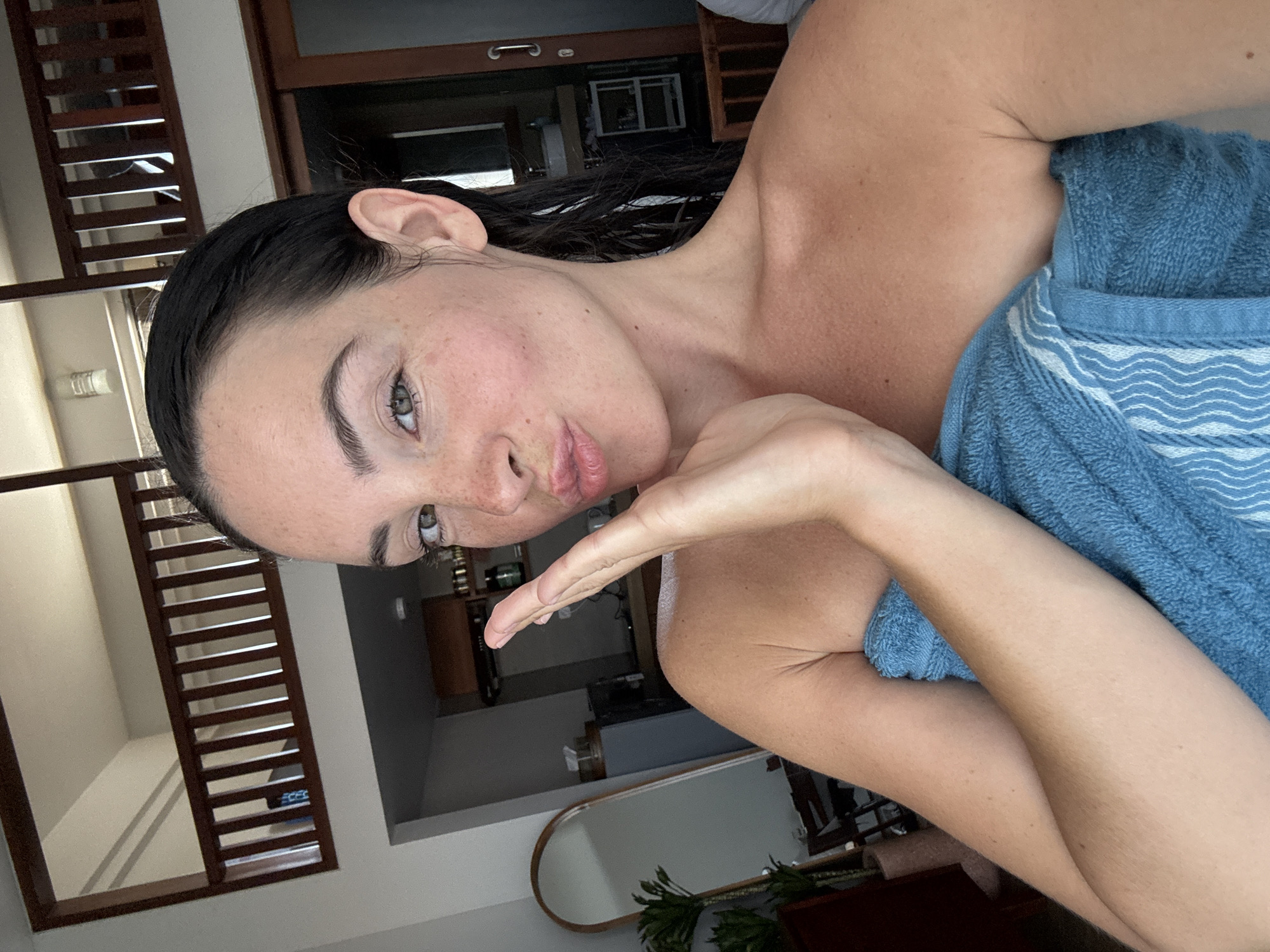 A woman with dark, wet hair wrapped in a blue towel poses for a selfie in what appears to be a bedroom or living space. She is making a kissing gesture with her lips. Her left arm is extended, with her hand resting on her cheek. The background includes a wooden railing on an upper level, a large arched mirror, some potted plants, and a doorway with sliding glass doors. The setting suggests a modern interior, possibly a loft-style apartment. The location is North Kuta, Indonesia.