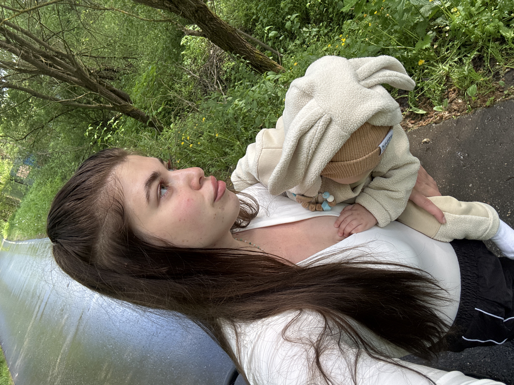 A young woman with long brown hair holds a baby wearing a beige bunny-ear hat and matching outfit. The woman is wearing a white top and black athletic shorts. They appear to be outdoors, possibly near a canal or waterway, given the glimpse of water and foliage in the background. The environment suggests a natural setting within a city. Without further details, it's impossible to confirm the location within Мещёры, Russia specifically.