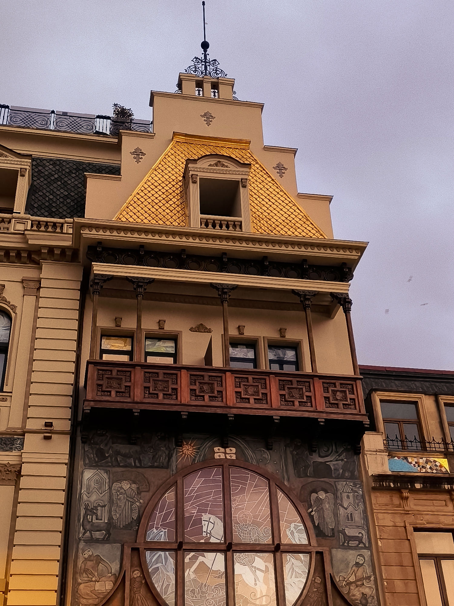 The image captures a detailed view of an ornate building facade in Batumi, Georgia. The architecture features a prominent golden, scale-patterned roof, intricate wooden balcony railings with carved details, and several windows. Below the balcony, a large, circular stained-glass window dominates the facade, with a mosaic-like patterned frame. The wall beneath the stained glass is adorned with a mural depicting various figures and scenes, including animals, what appears to be an angel, and human figures engaged in activities. The building's exterior is a warm, light brown color, with decorative moldings and columns visible. The sky above is overcast. To the right, a portion of an adjacent building with dark windows and a similar architectural style is visible.