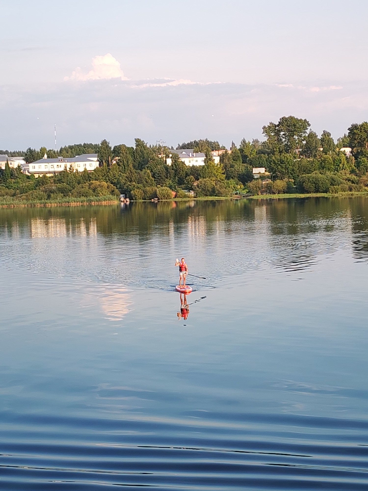 A person stands on a red paddleboard in a calm body of water, holding a paddle. The individual is wearing a life vest and appears to be waving. The water reflects the sky, trees, and buildings on the opposite bank. The background shows a shoreline with lush green trees and several white buildings. The sky is partly cloudy with a soft, diffused light. The scene is likely a tranquil summer day.