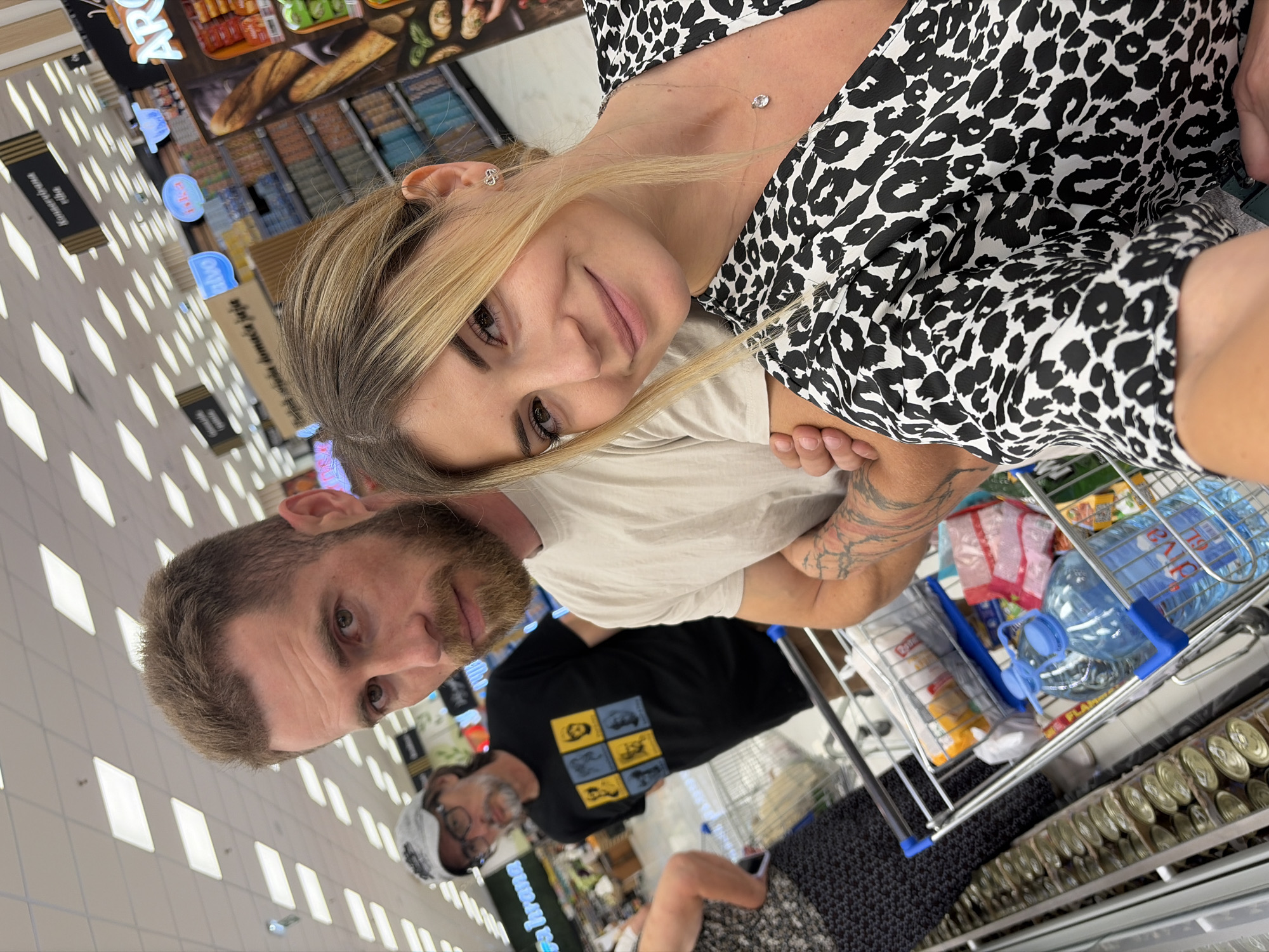 This is a selfie taken inside a supermarket in Budva, Montenegro. In the foreground, a young woman with blonde hair and a black and white leopard-print dress smiles at the camera, resting her head on the shoulder of a man beside her. The man, with short brown hair and a beard, wears a light-colored t-shirt and has visible tattoos on his arm. In the background, an older man wearing glasses and a grey baseball cap, and a woman in a patterned dress, are visible near a shopping cart filled with groceries, including bottles of water and canned goods. The supermarket aisle features shelves stocked with various products and overhead fluorescent lighting.