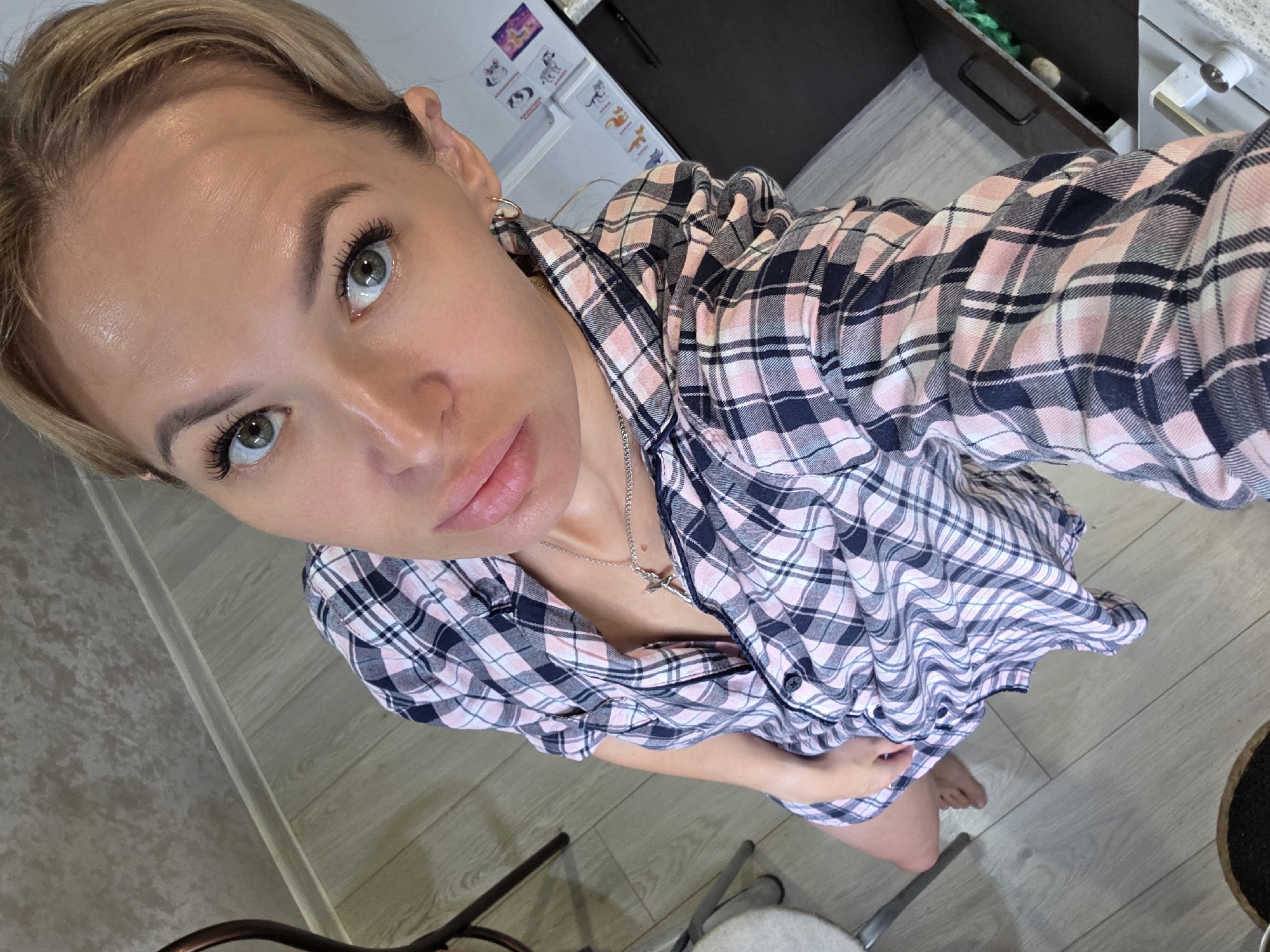 A blonde woman with green eyes and full lips, wearing a pink and blue plaid shirt and a silver cross necklace, poses for a selfie from a high angle. Her hair is short and styled in a bob. The woman is indoors, standing on a light-colored wooden floor. In the background, a dark cabinet or appliance is visible, as well as a chair and a small table. There are also some colorful stickers or drawings on the wall behind her. The image appears to be taken in a domestic setting.