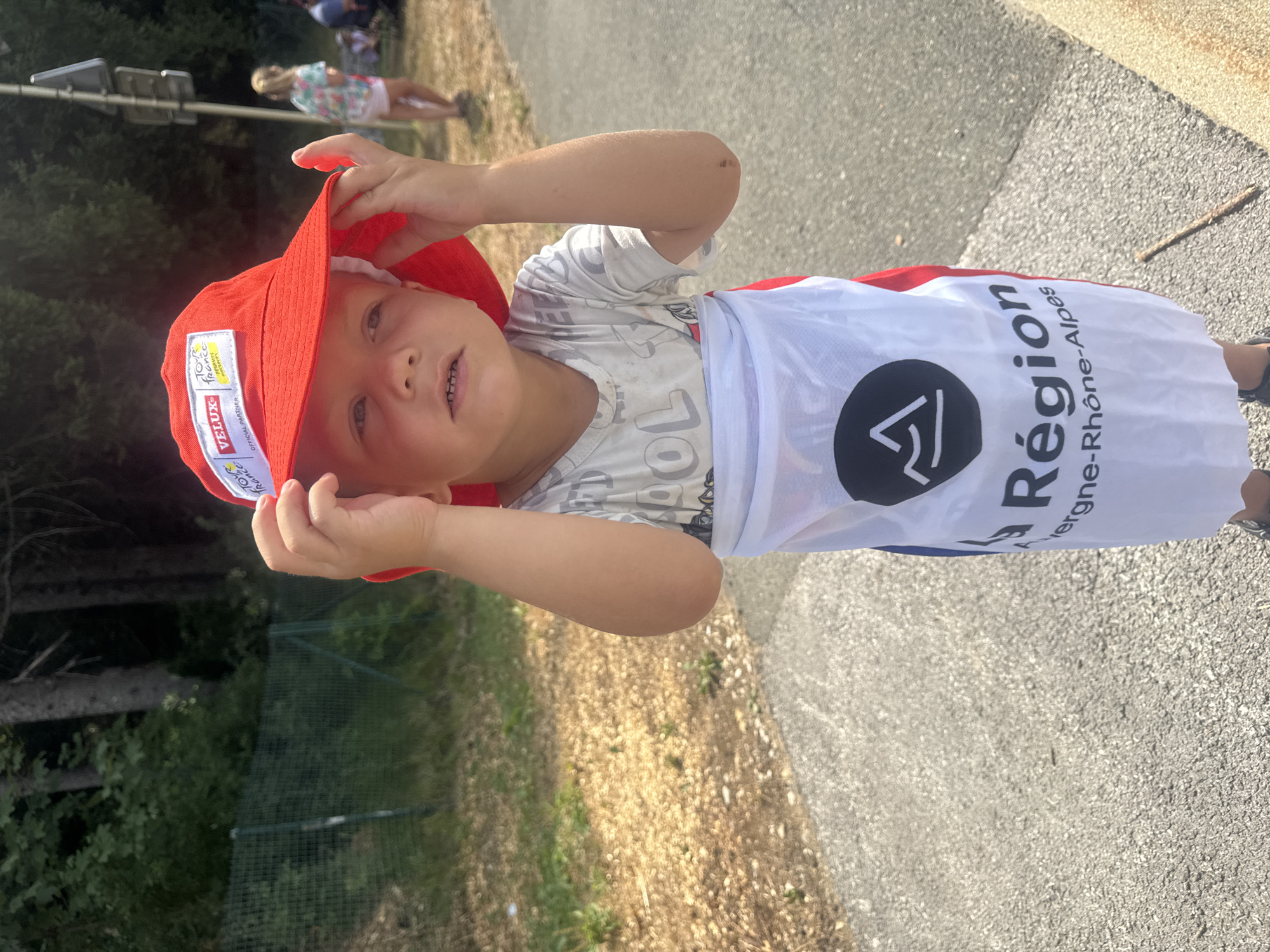 A young child, with fair skin and light hair, stands outdoors and is wearing a red bucket hat with the text "TOUR DE FRANCE" and "VELUX" visible on it, and a white garment with red accents that appears to be a cape or flag, displaying the words "La Région Auvergne-Rhône-Alpes" and a black mountain logo. The child is holding the brim of the hat with their right hand. The background shows trees, a fence, a paved path, and other people, including a girl in a floral print shirt and white shorts standing further in the background. The scene appears to be in a public outdoor setting, possibly a roadside event. The location is identified as Morbier, France.