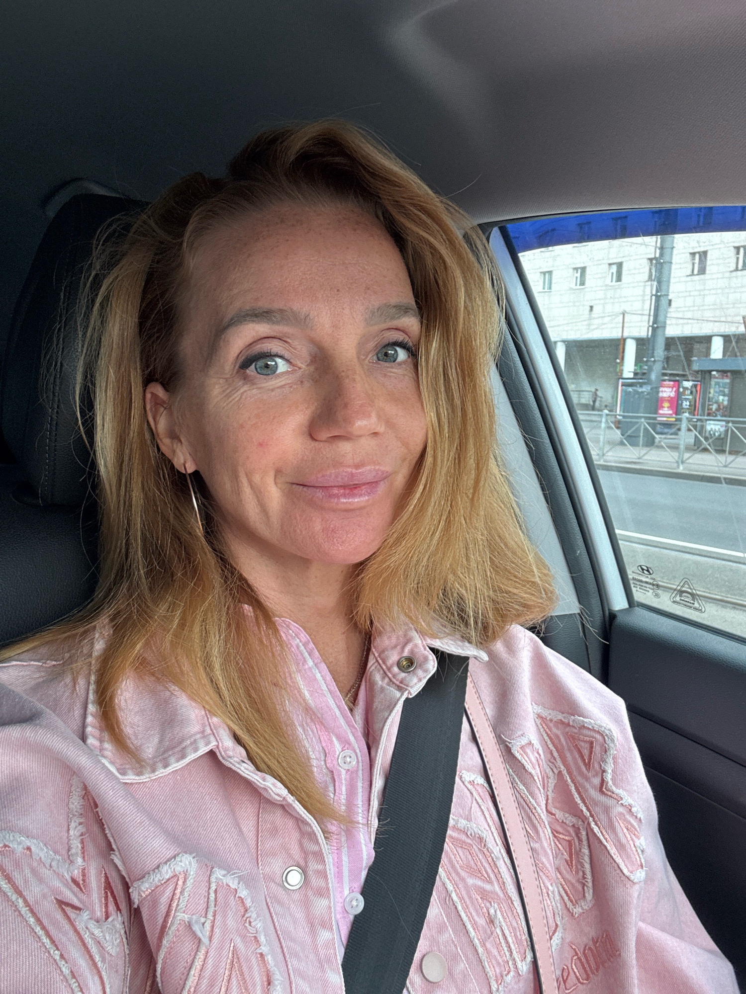 A medium shot portrait of a Caucasian woman with blonde hair and blue eyes, wearing a pink denim jacket over a pink and white striped shirt. She is in the driver's seat of a car, with a seatbelt fastened across her chest. Through the car window, a street scene in Saint Petersburg, Russia is visible, featuring buildings, streetlights, and tram tracks. The woman is looking directly at the camera with a slight smile. The overall lighting suggests an overcast or diffused daylight.