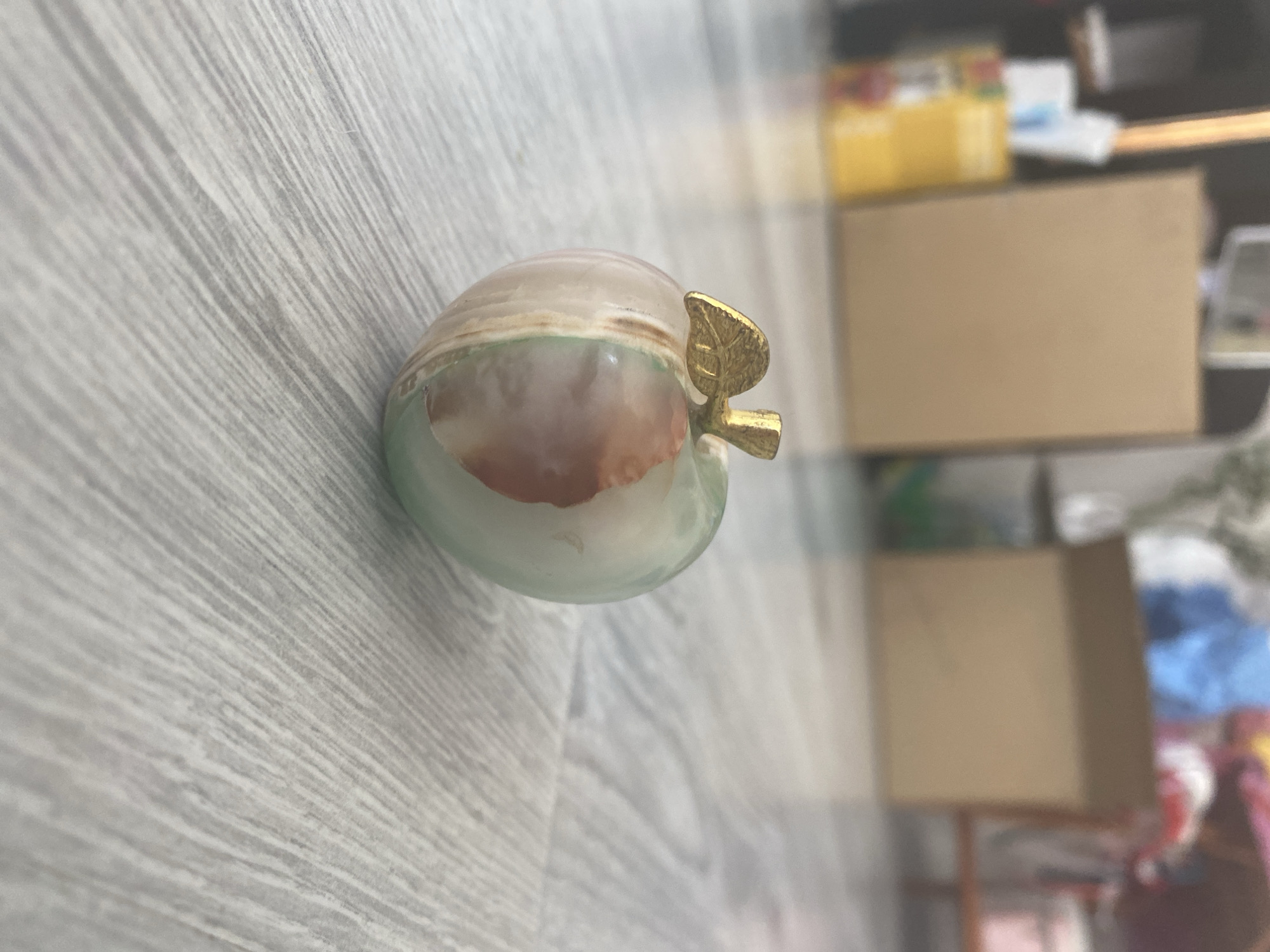 An ornamental apple figurine, crafted from what appears to be polished onyx with green and tan swirls, rests on a light-colored wooden floor. The figurine has a small, brass-colored leaf and stem. The background is blurred, showing cardboard boxes and other indistinct objects, suggesting an indoor setting possibly a storage area or a room undergoing organization. The lighting appears natural, casting soft shadows. The image does not provide any information about the city of Blagoevgrad, Bulgaria.