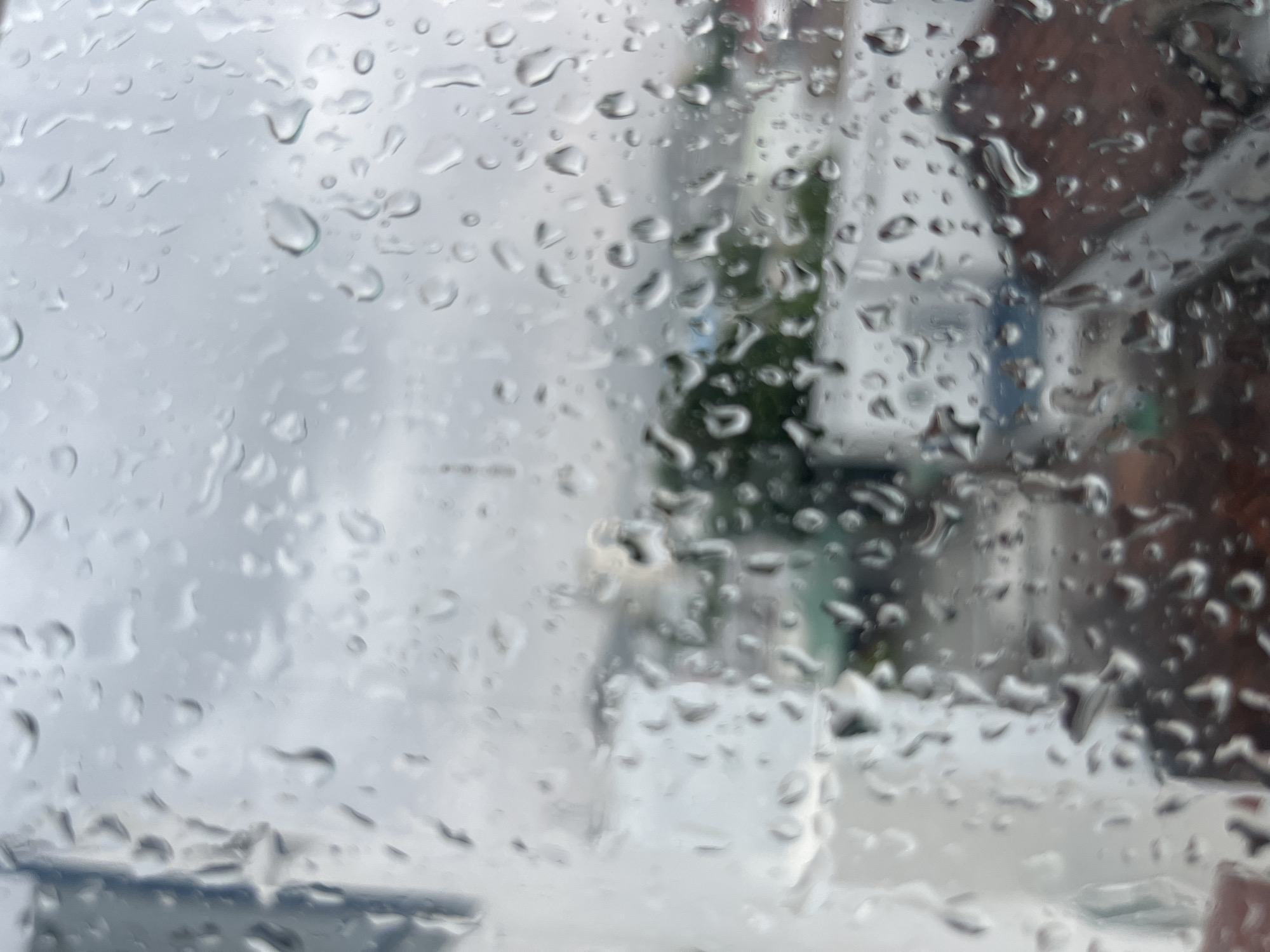 The image depicts a view through a rain-covered window. The rain droplets obscure the scene outside, but blurry shapes and colors suggest buildings, trees, and possibly cloudy sky. The scene is identified as being located in Bac Nha Trang Ward, Vietnam, but due to the distortion of the rain, details are limited.