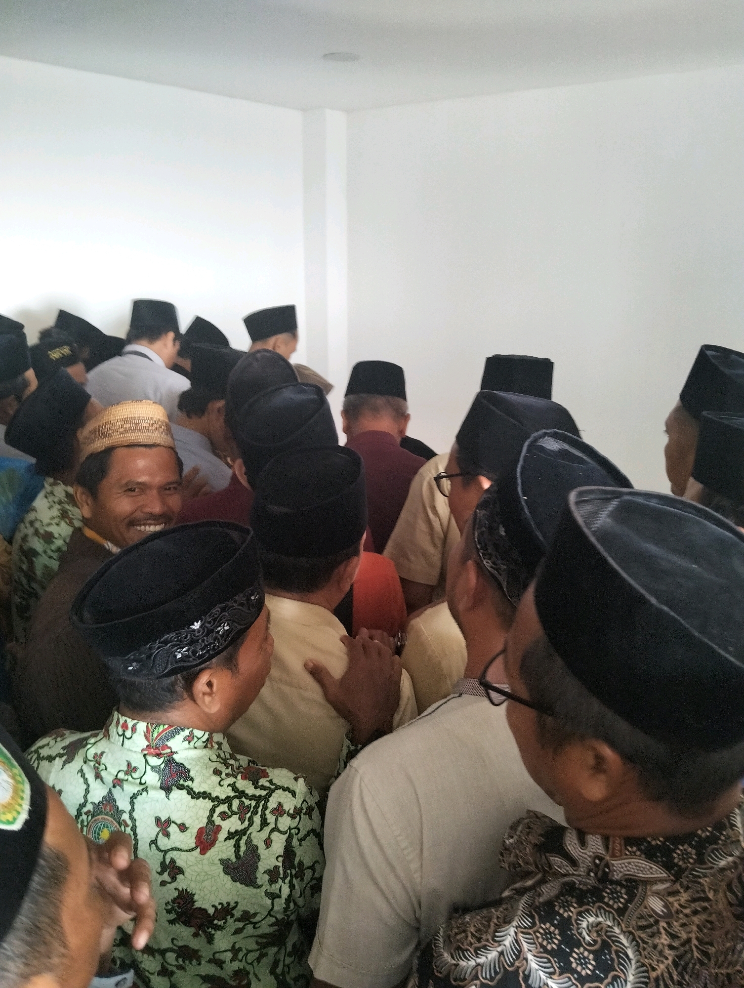 The image shows a group of men, most wearing black peci hats, gathered in a room with white walls. The man in the foreground, slightly to the left, is smiling and wearing a woven straw hat and a brown shirt. He is looking towards the camera. Another man in the lower right foreground is wearing a black peci with decorative embroidery and a light-colored shirt with a batik pattern. In the mid-ground, several men are seen from behind, some wearing light-colored shirts and others in darker attire. The environment appears to be indoors, with bright, diffused lighting. The gathering seems to be informal, possibly a social event or a meeting. This scene is located in the city of Paciran, Indonesia.
