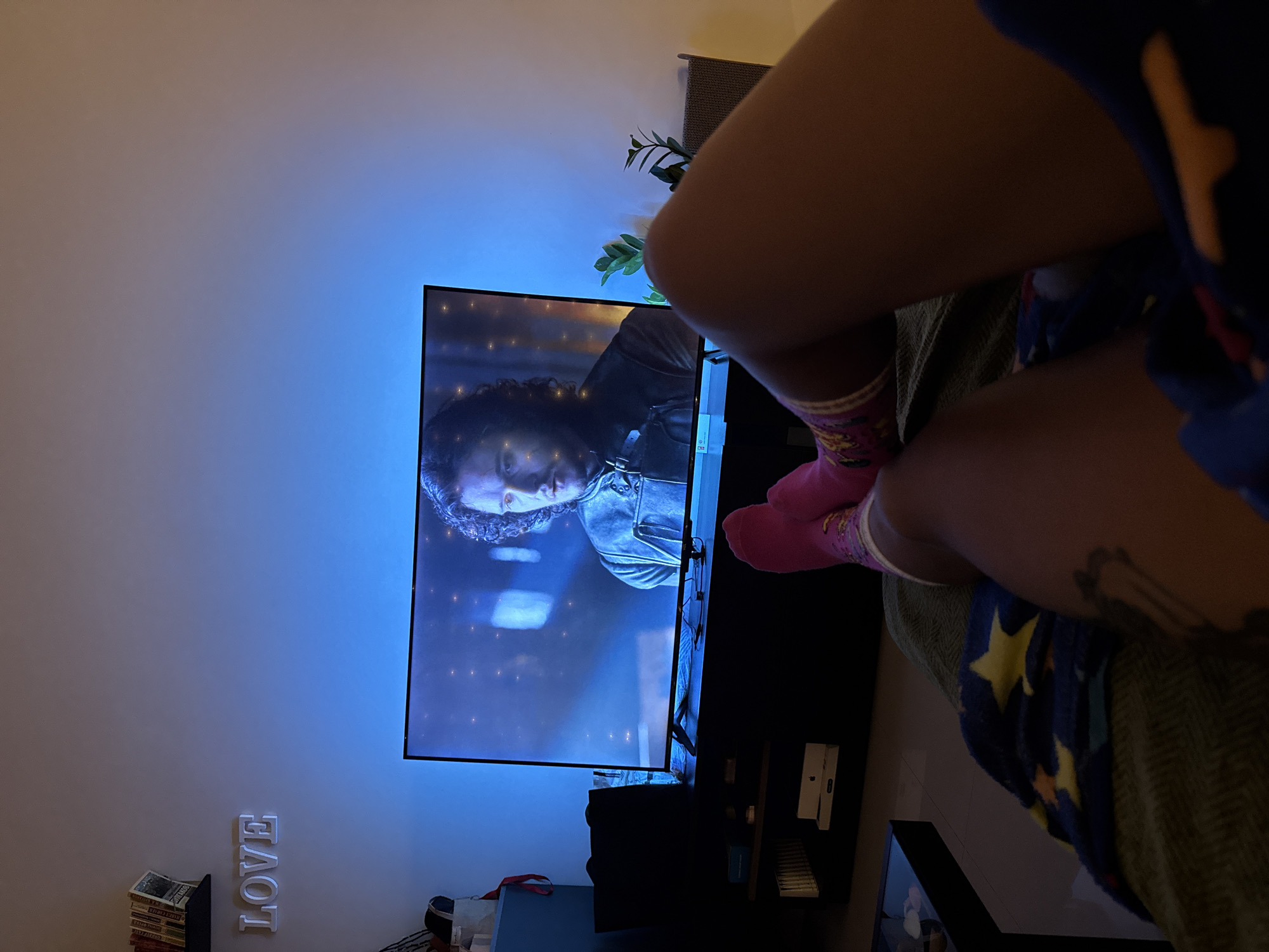 The image depicts a living room with a person lying on the floor, watching television. The room is dimly lit, with blue ambient light emanating from behind the television. On the wall above the television, the word "LOVE" is spelled out in white, three-dimensional letters, and a shelf with several books is visible to the left of the letters. The television screen displays a scene from a television show, featuring a man with curly dark hair wearing a leather jacket. In the foreground, the legs and feet of the person watching are visible, clad in pink and patterned socks. A blanket with a blue background and yellow stars is draped over the person. The scene appears to be within a residential setting in Thep Krasatti, Thailand.