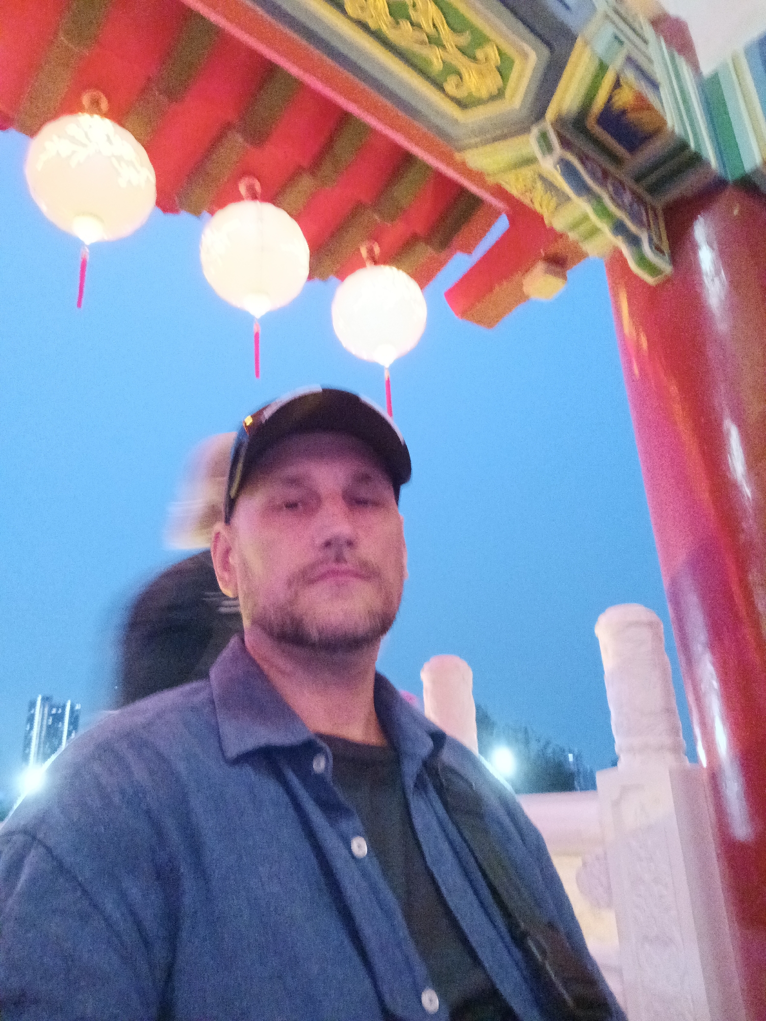 A man wearing a dark baseball cap and a blue button-up shirt is taking a selfie. He has stubble on his face and is looking towards the camera. Behind him, there are traditional Chinese lanterns hanging from a red and gold ornate structure. The sky is a clear blue, indicating it is daytime or twilight. The background shows glimpses of modern buildings and decorative white pillars, suggesting an urban setting that blends traditional and contemporary elements. The presence of the lanterns and architecture strongly suggests the location is within the vicinity of Kuala Lumpur's Chinatown or a cultural precinct.