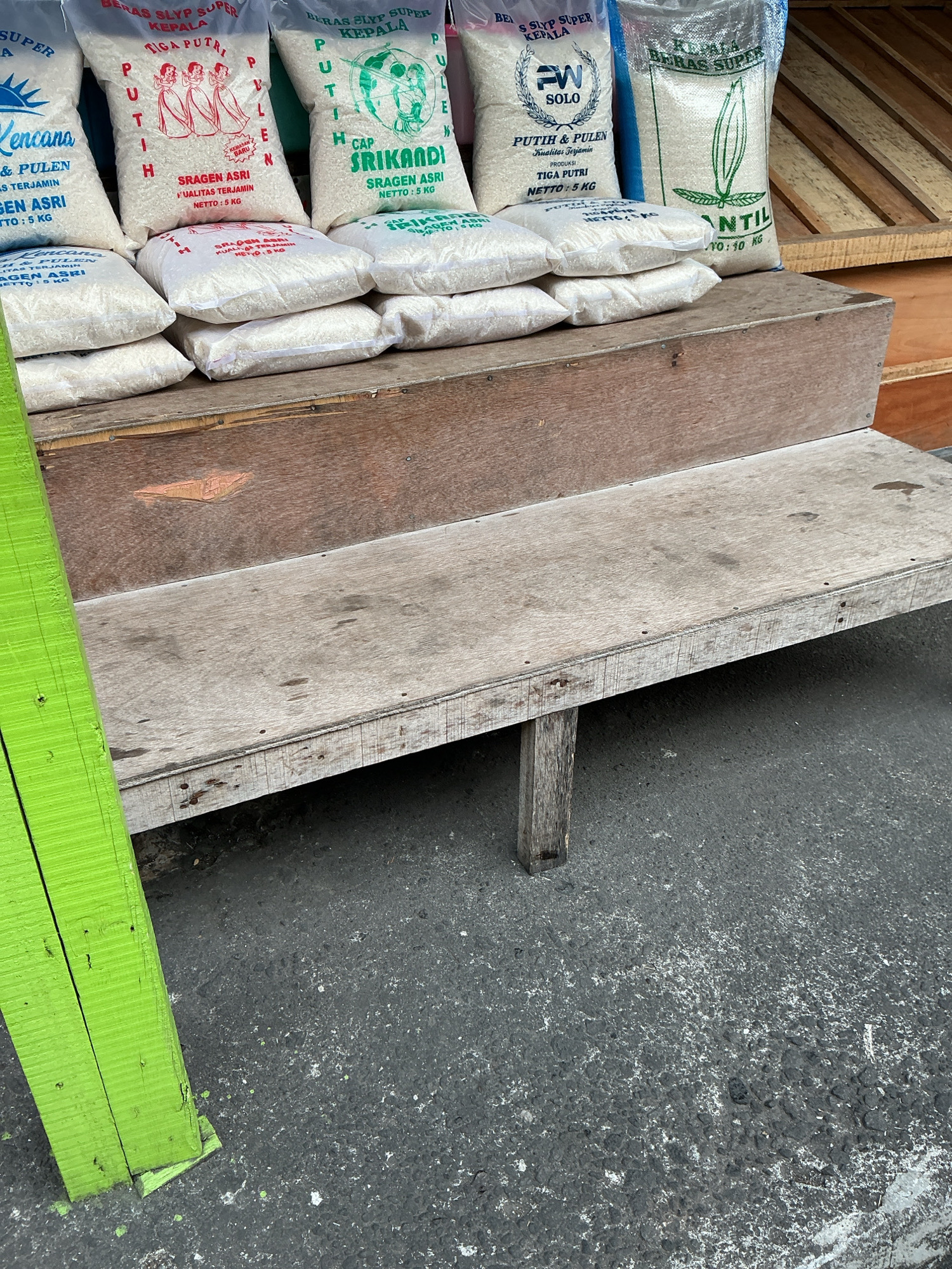 In the city of Magelang, Indonesia, an outdoor scene depicts a street-level view focusing on a display of rice sacks arranged on wooden tiered shelving. The rice sacks are primarily white with various labels indicating brands and weights, such as "BERAS SLYP SUPER KEPALA," "CAP SRIKANDI," and "ANTIL." Some sacks are stacked on wooden steps, while others rest on a wooden bench or platform in the foreground. The wooden structures appear weathered and worn. A bright green wooden plank stands vertically on the left side of the frame, leaning slightly into the scene. The ground surface is asphalt with some white speckling. The background shows more wooden structures, possibly part of a market stall or shop. There are no people visible in the image.