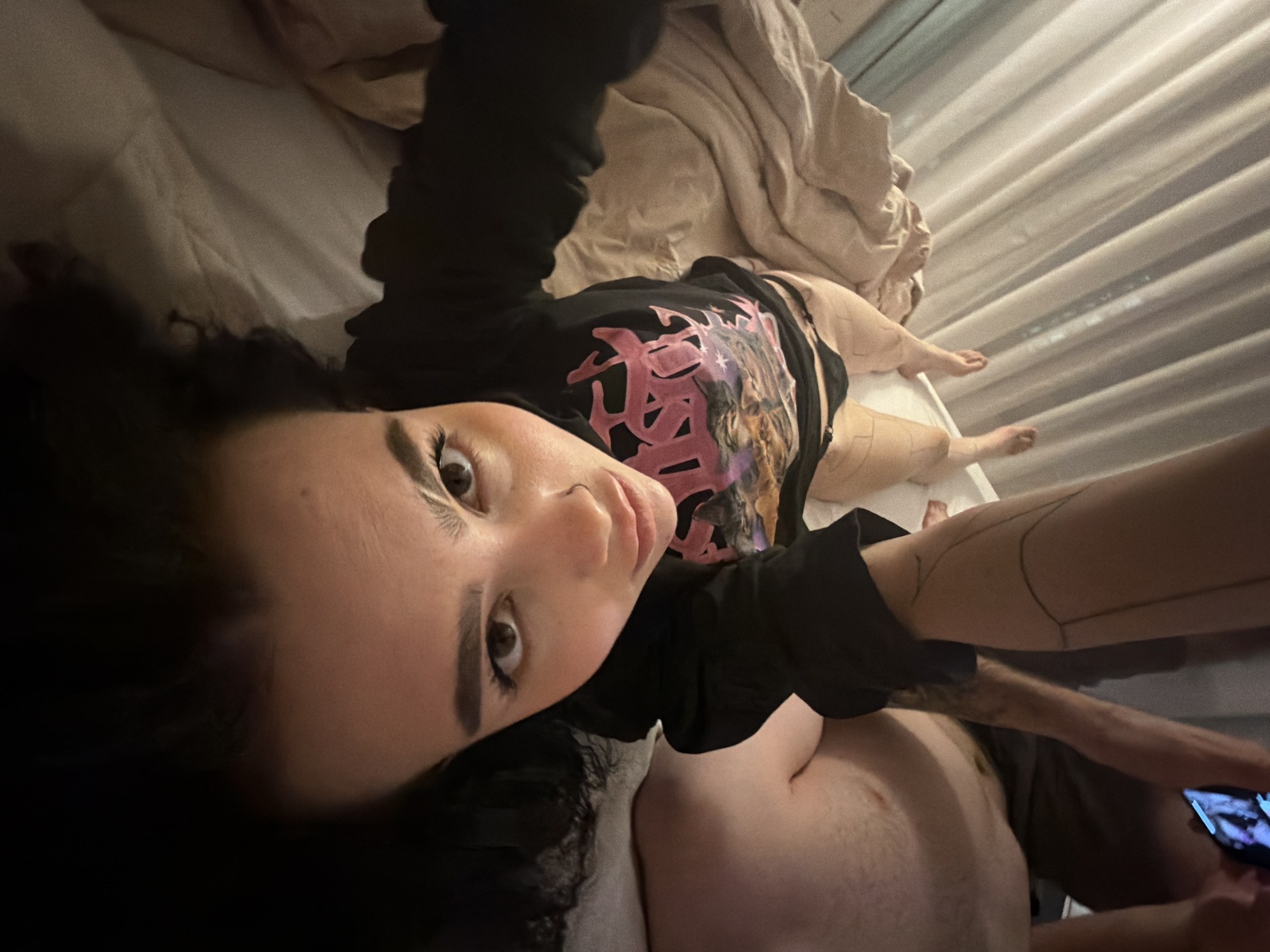 A selfie taken in an interior setting, likely a bedroom. A woman with dark curly hair is positioned in the foreground, lying on a bed. She is wearing a black long-sleeved shirt with a colorful design on the front. An unidentified individual, likely a man, is partially visible in the lower-right corner, with a bare chest and holding a smartphone. Bed sheets and a curtain can be seen in the background. There is no information to suggest the image was taken in Kazan, Russia.