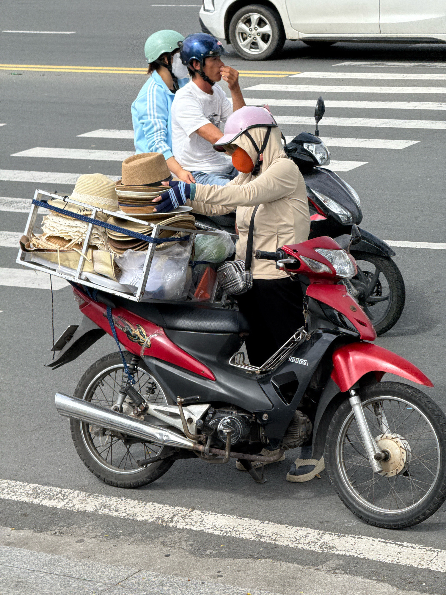 The image shows a street scene in Nha Trang, Vietnam, with a group of people on motorbikes stopped at a pedestrian crossing. In the foreground, a person wearing a beige hooded jacket and a pink helmet with an orange face covering is riding a red and black motorbike. The motorbike has a rack on the back carrying a large assortment of straw hats and some plastic bags. Behind this motorbike is a black motorbike with two people on it. The person in front is wearing a blue jacket and a light green helmet, while the person behind is wearing a white t-shirt and a blue helmet, holding something to their mouth. The road surface is asphalt with white pedestrian crossing lines. A white car is partially visible in the background.
