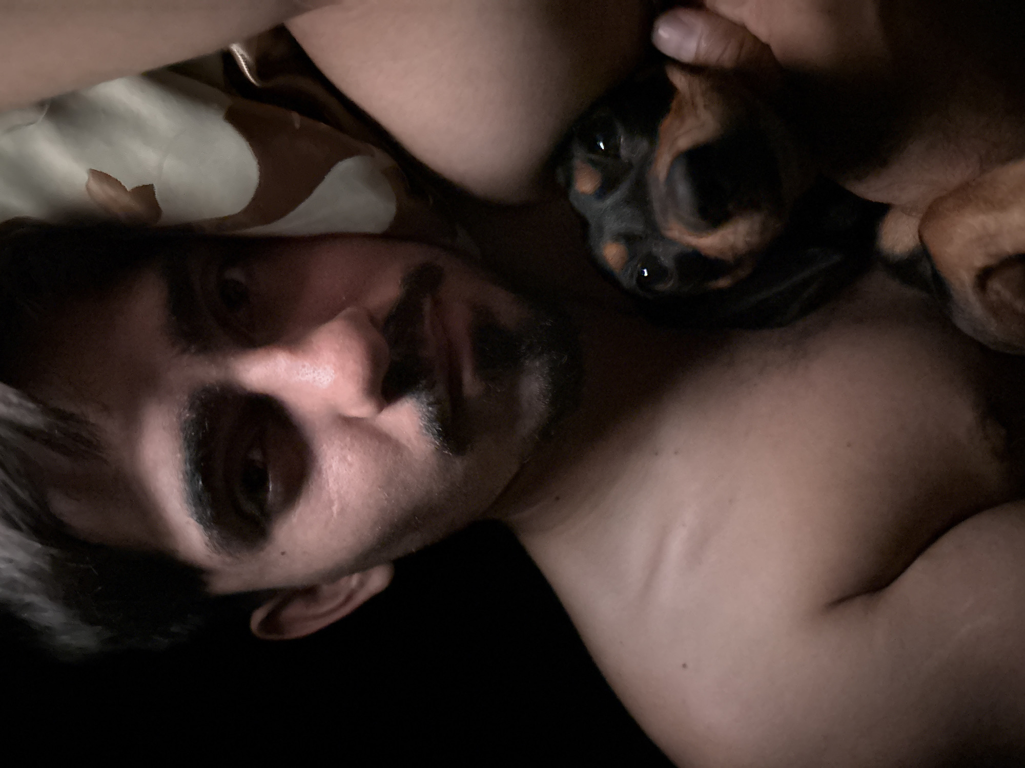 A shirtless man with dark hair and a mustache lies on a bed next to a black and tan dachshund puppy. The man's head is turned towards the camera, while the puppy is nestled against his body. A patterned pillow is visible in the upper left corner of the frame. The background is dark and indistinct. The location in the city of San Fernando del Valle de Catamarca, Argentina is not discernible from this image.