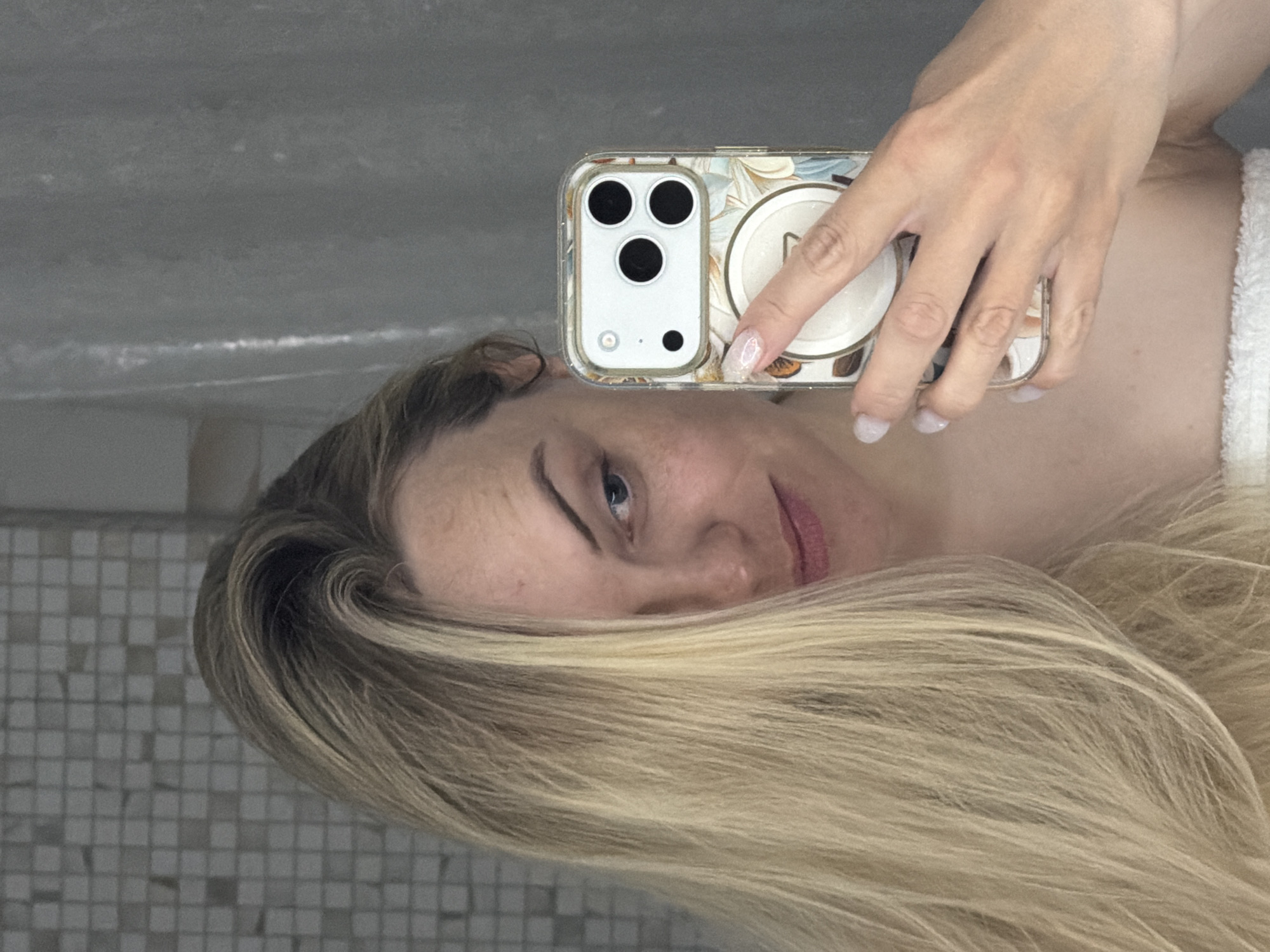 An indoor mirror selfie captures a person, likely female, from the chest up, with her long, flowing blonde hair cascading around her. The image is rotated 90 degrees clockwise, suggesting she might be lying down or reflecting in an angled mirror. She is holding an iPhone, which appears to be a newer model with three camera lenses, in a clear case adorned with a delicate floral pattern, possibly featuring light-colored flowers. A white pop socket with a metallic-looking "N" or similar emblem is attached to the back of the phone. Her hand, with manicured nails painted in a light, possibly glittery, polish, holds the phone. A white towel is visible around her shoulder or neck.
The subject has a neutral to slightly contemplative expression, looking towards her reflection. Her visible eye is blue. The background reflected in the mirror shows a tiled wall on the lower left, characterized by small, square, light-colored tiles, and a plain, light gray surface, possibly a ceiling or upper wall, on the right. The setting suggests a bathroom, spa, or changing room. The overall context for this image is Votkinsk, Russia.
