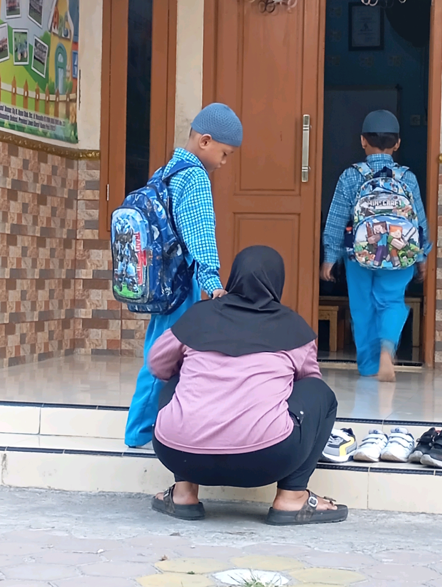 The image depicts an outdoor scene in front of a building with brown double doors, likely in Babelan, Indonesia. Three individuals are present. On the left, a young boy wearing a blue skullcap and a blue checkered shirt with a backpack featuring images of robots is seen from the side, looking down. In the center, another young boy, also wearing a blue skullcap and a blue checkered shirt, carries a backpack with "Minecraft" characters and is walking away from the viewer towards the open doors. A third person, dressed in a purple hijab and a black head covering, is crouched down in front of a row of shoes on a stepped platform. The shoes include a pair of grey and yellow sneakers, a pair of white sneakers, and a pair of black sneakers. The ground is paved with light-colored square tiles. The building's facade features a patterned brick wall on the left, and a colorful mural with depictions of a house and a fence is visible on the upper left. The lighting suggests it is daytime.