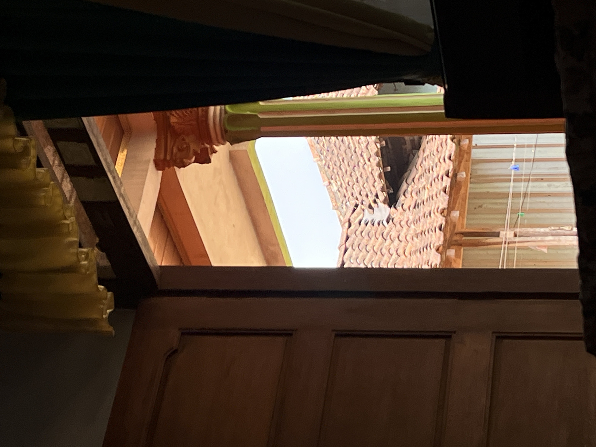 The image depicts a view from inside a wooden doorway looking out onto a tiled roof structure. On the left, a portion of a closed wooden door is visible, appearing to be made of dark wood with distinct paneling. The doorway opens to an interior space with an orange ceiling and a wooden beam. To the right, a decorative pillar with orange, green, and pink stripes supports the ceiling. Beyond the pillar and the doorway's frame, a traditional Indonesian tiled roof with terracotta tiles is prominent. The background shows a light-colored sky and hints of vertical striped curtains or walls. There are no people or animals visible in the scene. The overall impression is of an outdoor view seen through an open doorway, with architectural elements of a building in Glagah Kulon, Indonesia.