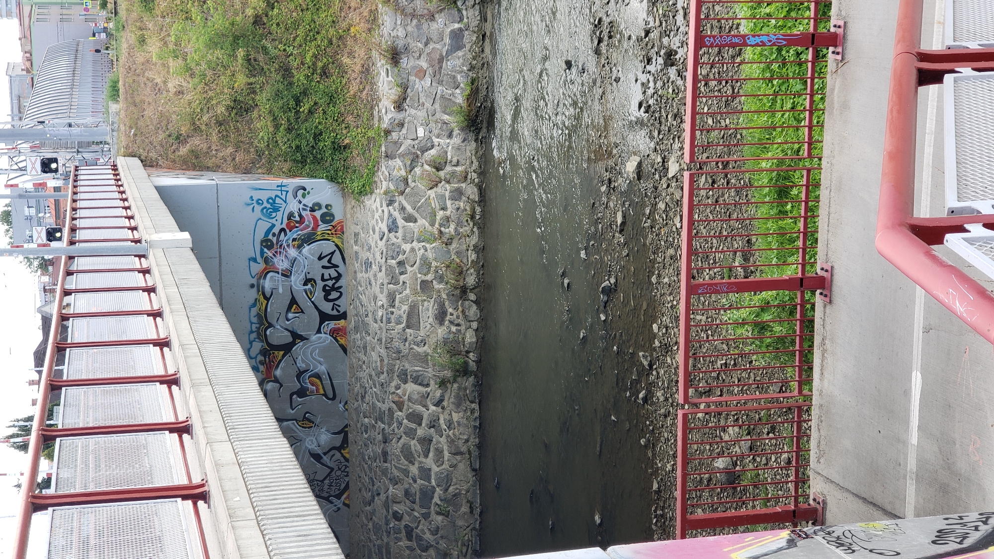 The image captures a view of a bridge over a small river or stream in Prešov, Slovakia. The bridge has a concrete deck supported by concrete pillars. The underside of the bridge is adorned with graffiti, with the word "CREW" being clearly visible. The riverbed is rocky and the water appears somewhat murky. Red railings line the sides of the bridge and a lower walkway. Vegetation, including grass and some small bushes, grows on the embankment behind the railings. In the background, there are buildings, some industrial in appearance with curved roofs, and what appear to be railway crossing signals. The sky is overcast.