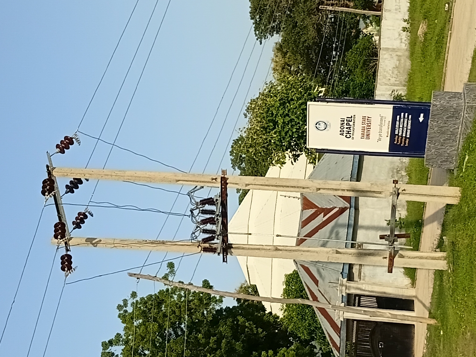 A daylight, medium shot captures a street scene in Jalingo, Nigeria, focusing on utility poles and a sign. In the foreground, two large, pale concrete utility poles stand prominently, supporting multiple black electrical wires suspended by dark brown ceramic insulators. The wires stretch across the frame, disappearing into the background. Between the poles, a weathered, grey metal crossbar is attached to the left pole. To the right of the utility poles, a white rectangular sign with a dark blue base stands upright. The sign displays text in dark blue and grey font, including "ADONAI CHAPEL OF TRANSFORMATION" and "TARABA STATE UNIVERSITY." Below this, a slogan reads, "Be ye transformed" and lists weekly activities. A white arrow points to the left at the bottom of the sign.
In the midground, behind the utility poles, a white building with a conical, silver-colored roof is visible, partially obscured by trees. To the left of this building, a white wall with decorative pillars and a dark metal gate with vertical bars is present. The roof of a smaller structure in front of the white building has a corrugated metal surface with alternating red and brown stripes. Lush green trees and grass cover the surrounding landscape, suggesting a well-maintained environment. The sky above is a clear, light blue. The scene appears calm and depicts an outdoor urban setting with infrastructure and signage.