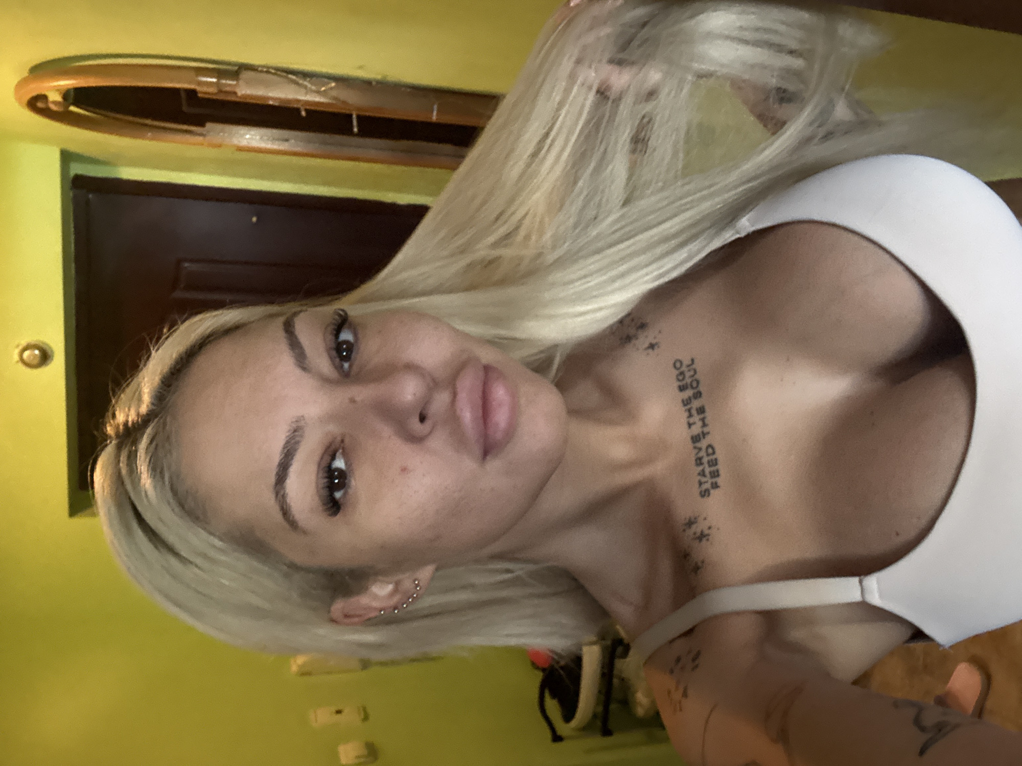 A blond woman is posing for a selfie. She is wearing a white top, and she has multiple piercings in her left ear. She has tattoos on her chest and arms. The background shows a green wall and a brown door. It is not possible to determine if this photo was taken in the city of Plovdiv, Bulgaria.