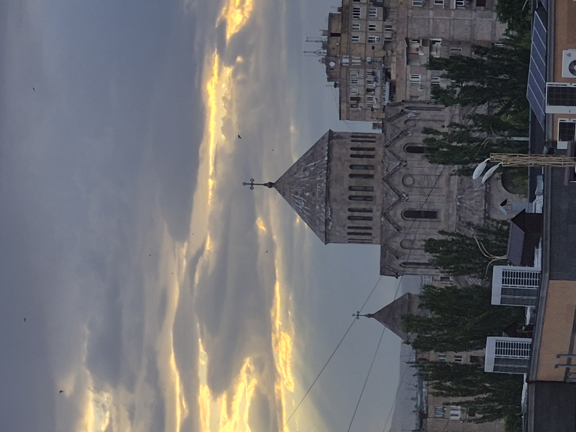 The image displays a cityscape in Yerevan, Armenia, captured during either sunrise or sunset, presenting a dramatic sky above urban architecture. The upper portion of the frame is dominated by a striking sky where deep blue-grey clouds are intensely illuminated by golden and yellow light from the low-hanging sun, creating a vibrant and contrasting display. Several small, dark specks, likely birds, are visible soaring through the illuminated sky.
In the midground, a distinctive Armenian Apostolic church or cathedral stands prominently. It is constructed from light-colored stone and features a multi-tiered, pointed roof, topped with a cross. The church's facade showcases arched windows and intricate architectural detailing. A smaller, similar spire with a cross is partially visible further back.
To the right foreground, the upper section of a modern building is visible, equipped with several white, rectangular air conditioning units and angled solar panels on its roof or side. In the background, behind the church, multiple multi-story residential apartment buildings rise, characteristic of Soviet-era architecture, with their beige facades and numerous windows. Green foliage from trees is interspersed throughout the urban landscape, adding a natural element among the structures. No specific activities beyond birds flying are depicted, and no visible text is present.