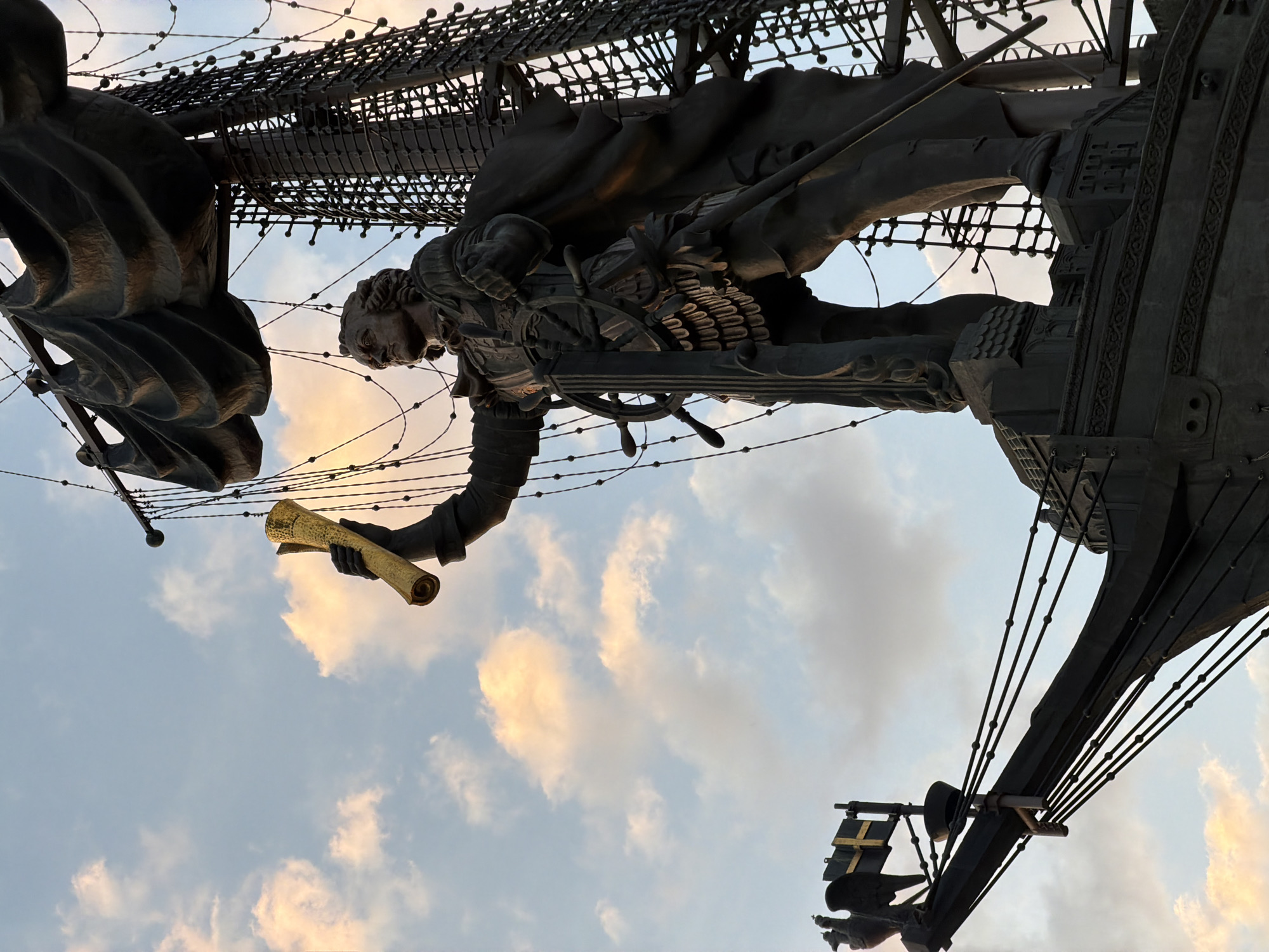 The image is a low-angle shot of the Peter the Great Statue in Moscow, Russia, against a backdrop of a cloudy sky with a hint of sunset colors. The statue depicts Peter the Great holding a scroll in his outstretched hand, standing on what appears to be a ship. The statue is made of dark metal, possibly bronze or steel, and has intricate details such as the rigging of the ship and the design of Peter's clothing. The statue appears to be quite large.