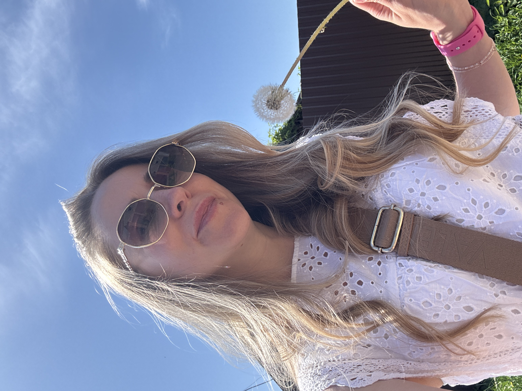 A woman with long blonde hair wearing sunglasses and a white eyelet top is holding a dandelion. She has a pink watch and a bracelet on her right wrist. A brown bag strap crosses her torso. The background shows a blue sky with clouds and a dark brown building. The location is Chaikovsky, Russia.