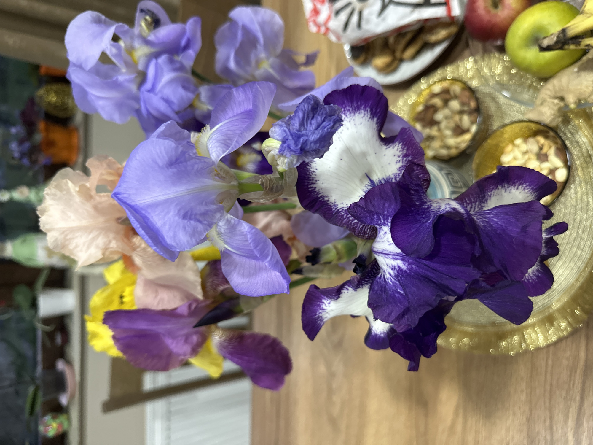The image shows a still life arrangement on a wooden surface. A vase containing irises in various colors, including blue, purple, white, yellow, and peach, is the central focus. Adjacent to the flowers are snacks and fruits, including a plate with cookies, apples, and a banana. A golden platter containing nuts is also present. The image's location is not discernible based solely on the provided information.