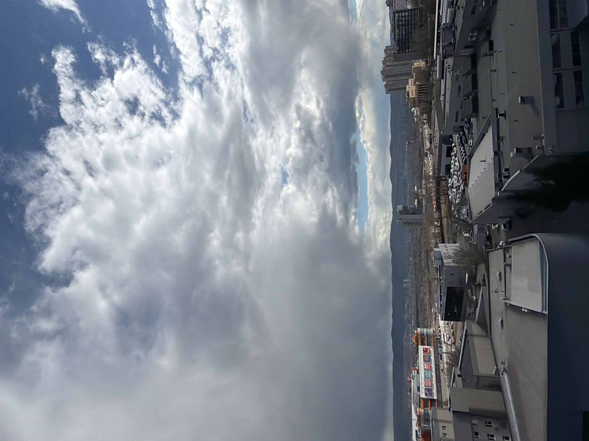 This elevated panoramic view captures a cityscape in Krasnoyarsk, Russia, under a partially cloudy sky. The sky dominates the upper portion of the image, displaying a dynamic mix of weather conditions: patches of clear blue sky are interspersed with white, fluffy cumulus clouds on the left, transitioning to a thicker expanse of uniform grey clouds on the right.
In the distance, a long chain of muted mountains or hills forms the horizon line, separating the sky from the urban development below. The cityscape itself features a blend of multi-story buildings, including what appear to be residential towers and various commercial or industrial structures with flat rooftops. A large parking lot filled with numerous vehicles is visible among the buildings.
The immediate foreground reveals parts of modern building rooftops, predominantly grey and industrial in appearance, possibly belonging to commercial complexes. The lighting suggests it is daytime, likely mid-morning or afternoon, with soft, diffused sunlight. There are no identifiable people or specific activities taking place, presenting a static urban landscape. On an orange building in the mid-ground, a white sign with text is visible, but the words are unreadable due to the distance and resolution.