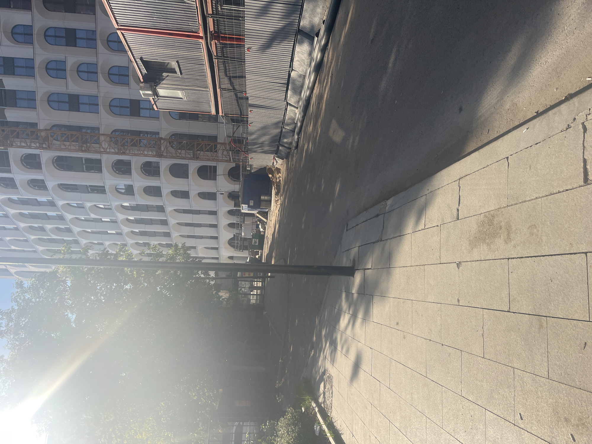 The image shows a construction site with a large, modern building under construction in the background. The building has a facade with numerous arched windows. A construction crane is visible next to the building. In the foreground, there is a sidewalk made of light-colored paving stones, and a road. Construction barriers and temporary structures are present along the side of the road. The scene is lit by strong sunlight, casting shadows. The image appears to be taken in Moscow, Russia, based on the architectural style and construction practices commonly seen in the city. There are no people visible in the image.