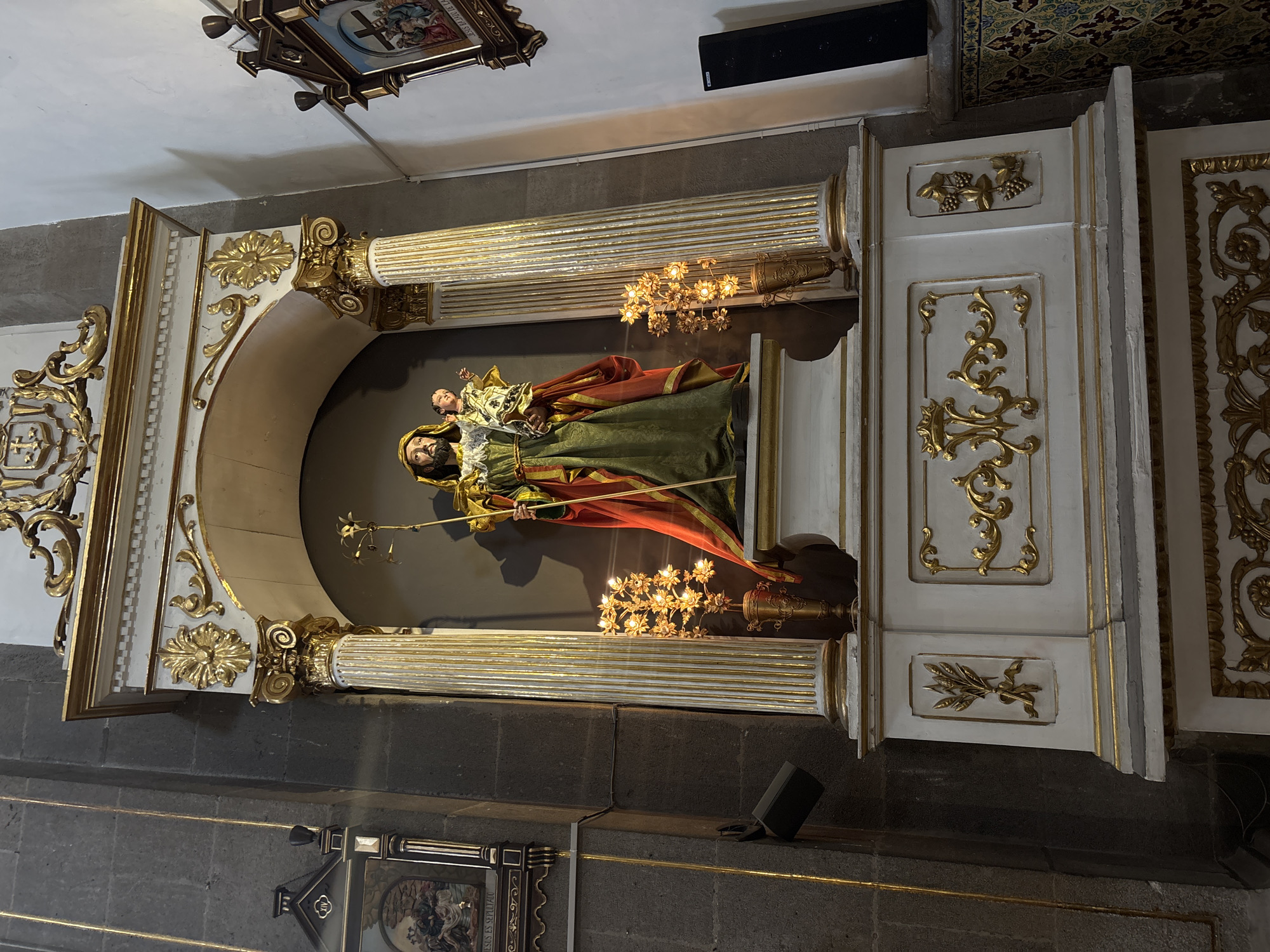 The image depicts a religious statue of Saint Joseph holding the infant Jesus, placed within an ornate alcove inside a church. The location is the Iglesia de Santo Domingo (Templo de Santo Domingo), Puebla, Mexico.
The statue is centered in a niche decorated with gilded elements and Corinthian-style columns. Below, candlesticks holding multiple small candles flank the statue. The alcove structure is white with gold ornamentation, extending outward from the dark gray stone wall of the church. A framed picture is visible above the statue. Another framed painting is partially visible below the statue.