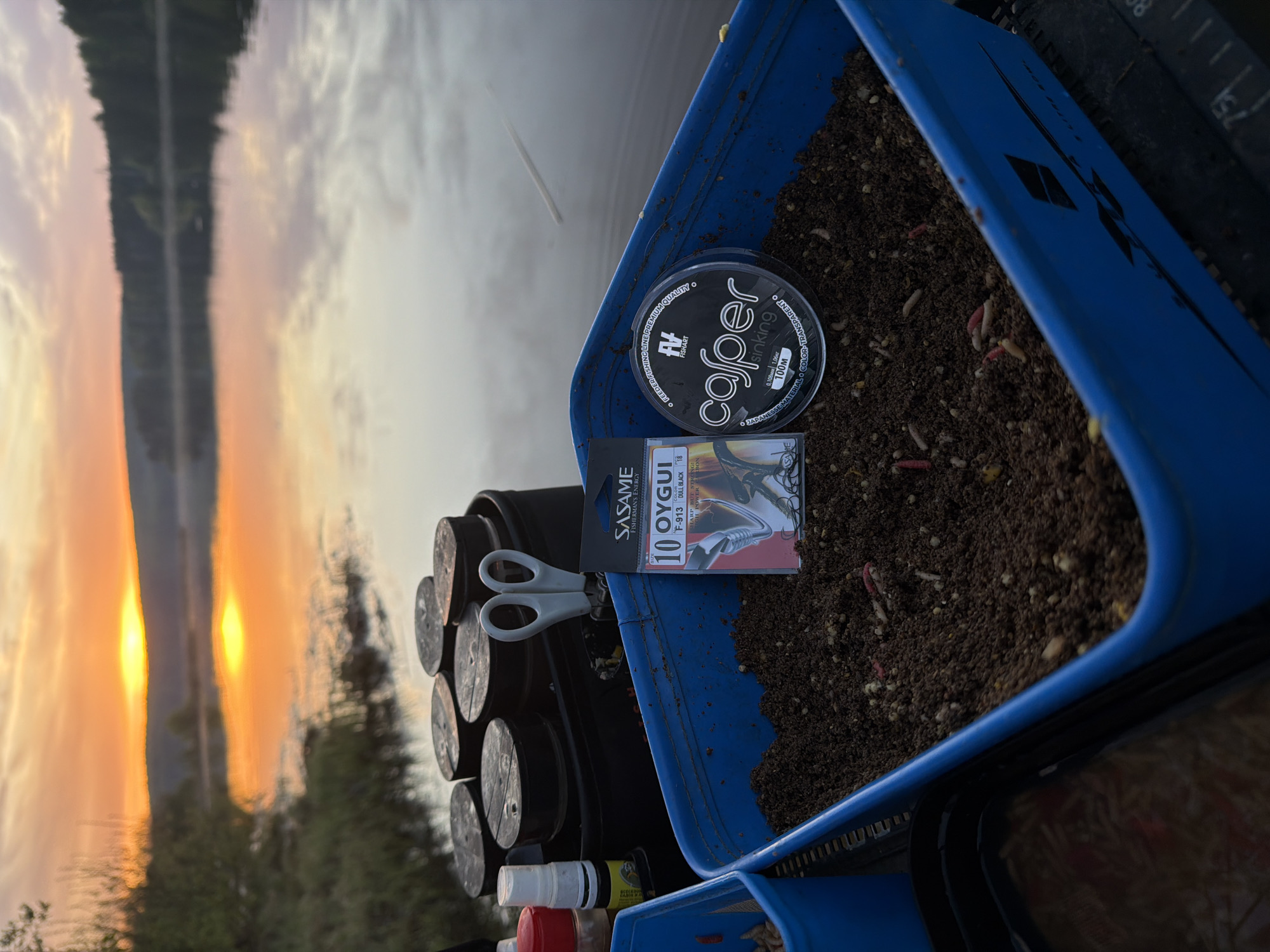 The image shows a fishing setup beside a body of water with a sunset reflection, possibly taken in the city of Chaykovsky, Russia. The foreground features a blue container holding dark soil and bait, including worms and larvae. On top of the container are fishing-related items: a spool of "casper" brand fishing line, a package of "SASAME OYGUI" hooks, and a pair of scissors. Stacked beside the container are multiple spools of fishing line. Partially visible below the container are bottles or containers with red, white, and yellow caps. In the background, the setting sun reflects off the water, creating a bright orange glow. Silhouetted trees or land are visible on the opposite shore.